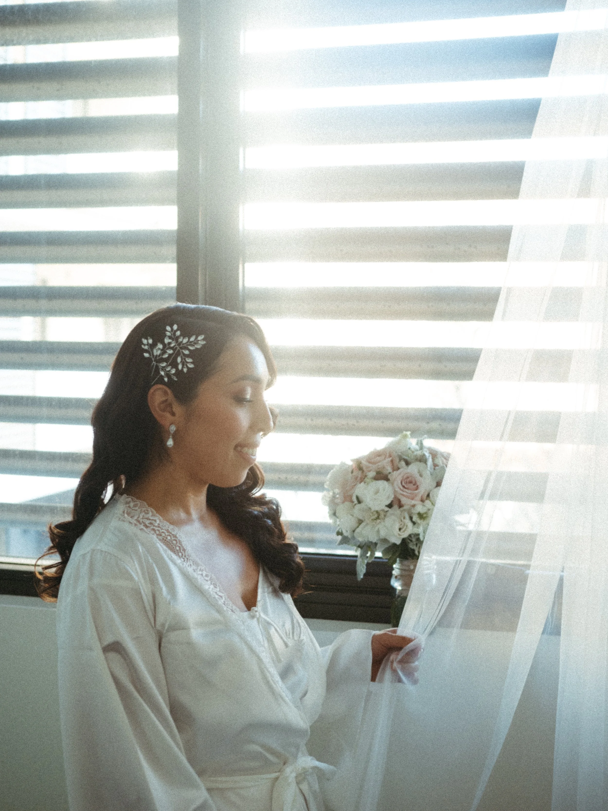 A woman with dark hair wearing a white robe, holding sheet music, standing next to a bouquet of white and pink roses, indoors near a window with horizontal blinds.
