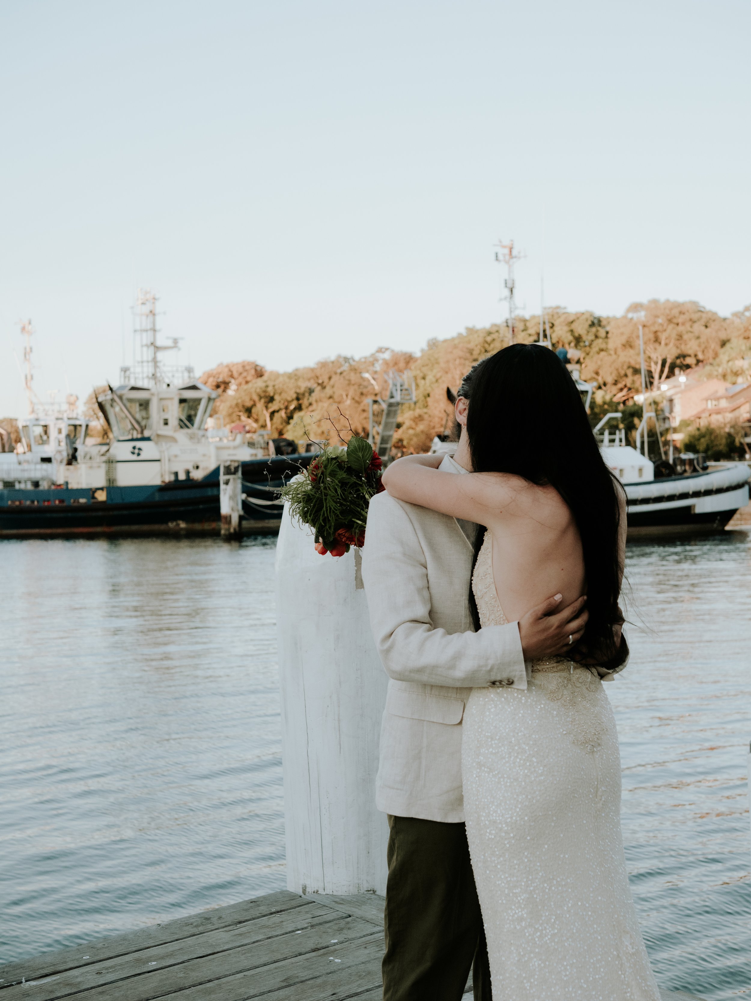 A couple embracing by the waterfront during sunset, with boats and trees in the background.