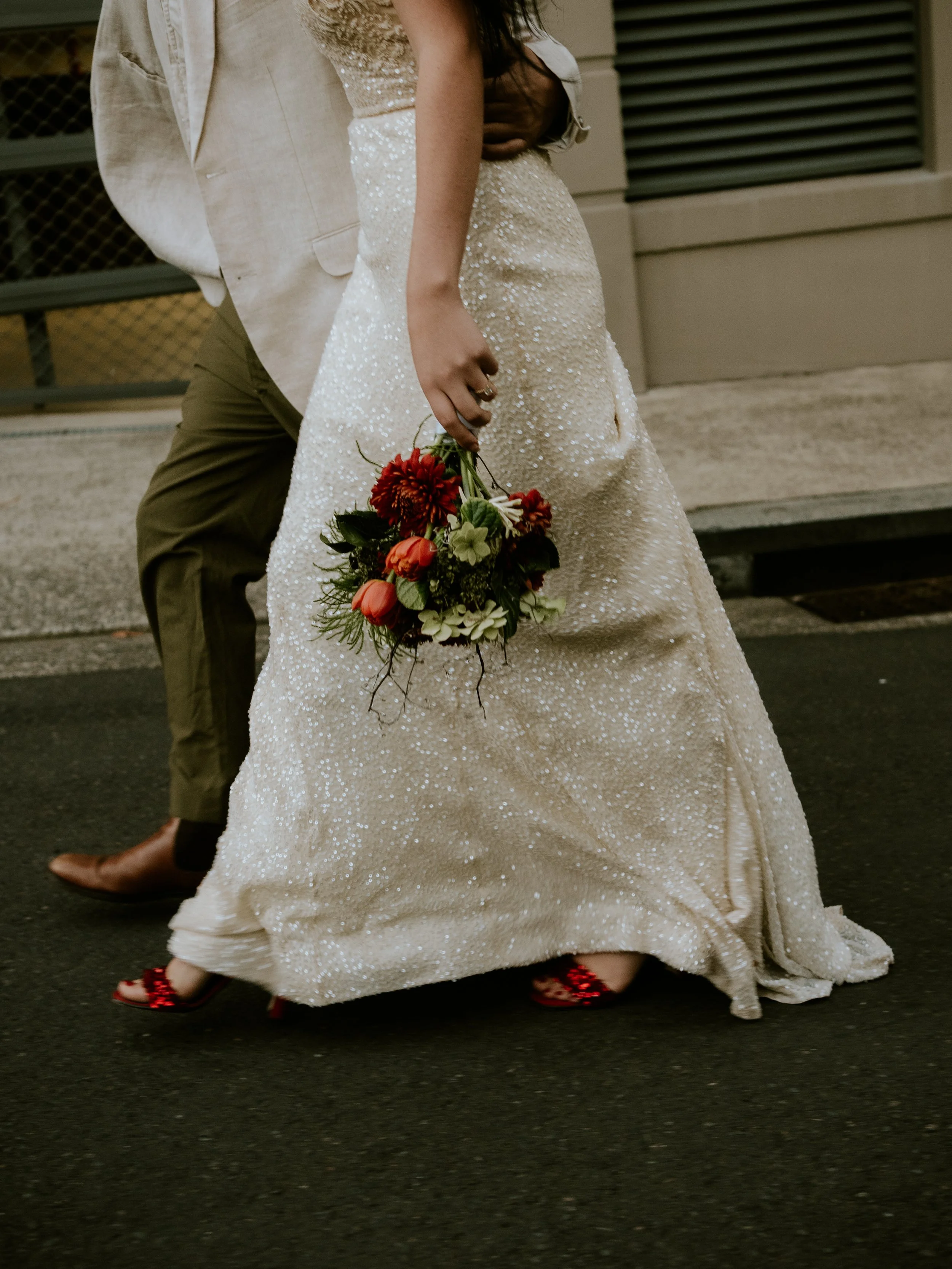 A bride in a sparkling white wedding gown holding a bouquet of red, orange, and green flowers, with a person in a beige jacket and olive green pants walking beside her on a city street.