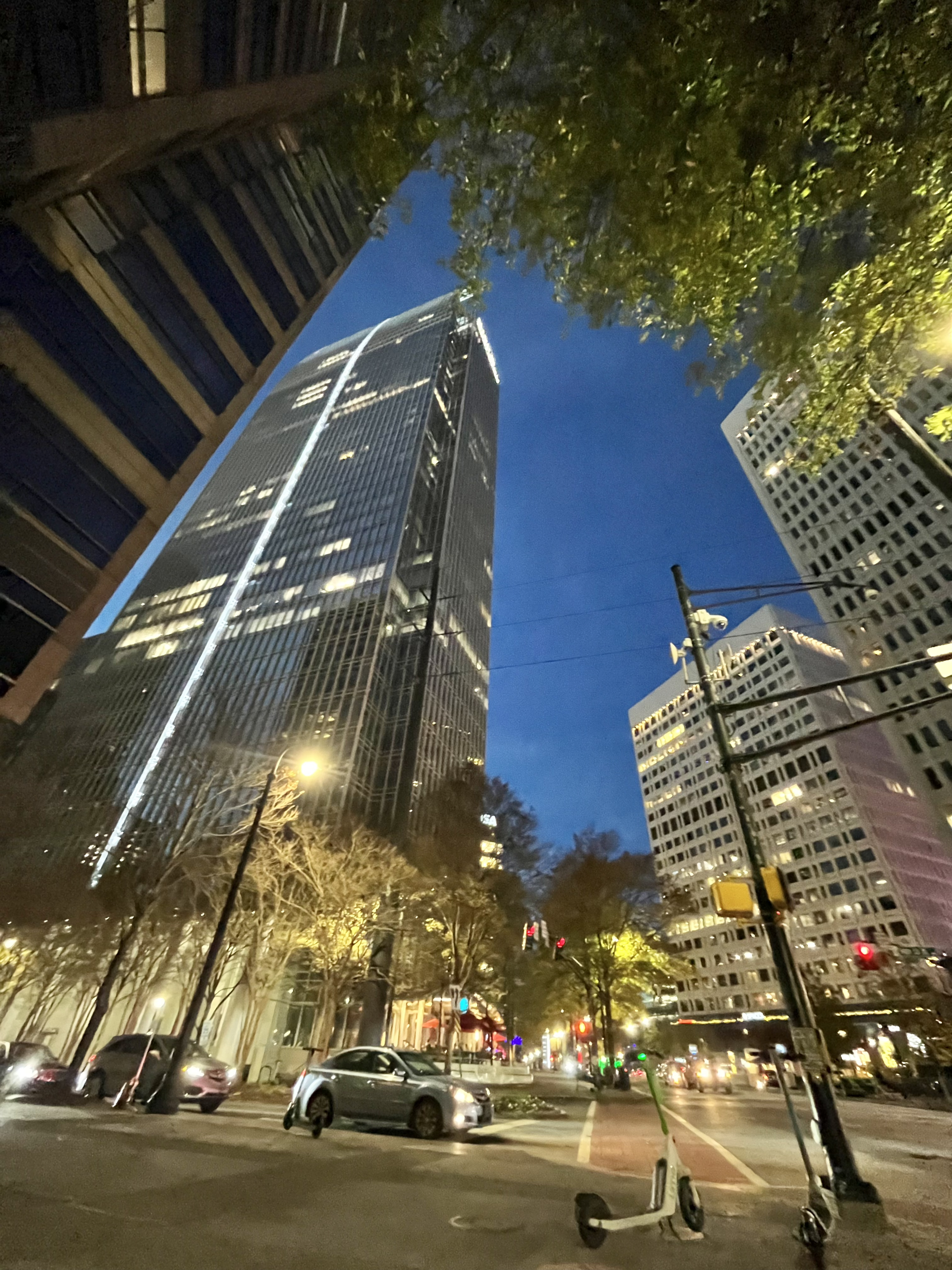 A nighttime city street scene with tall skyscrapers, trees, parked cars, and lit streetlights. Several electric scooters are parked in the foreground, and the sky is dark blue.