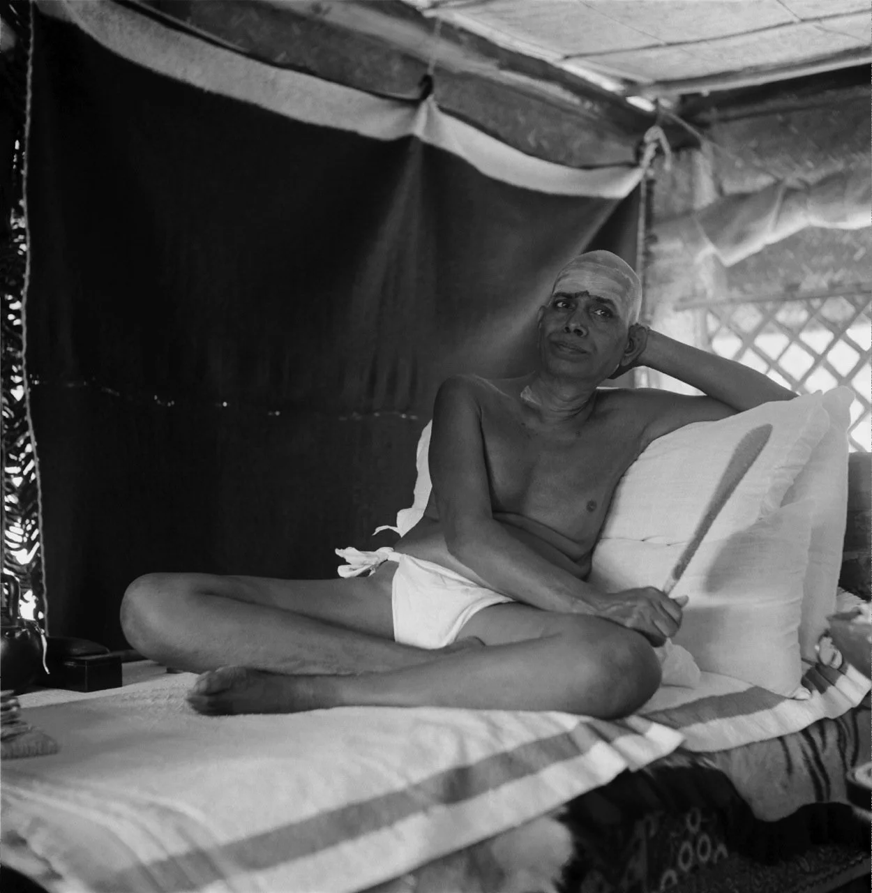 An elderly man with a shaved head, sitting on a bed with a pillow, inside a simple room with a wooden roof and a lattice window in the background. He is shirtless, wearing shorts, and holding a stick, resting his head on his left hand.