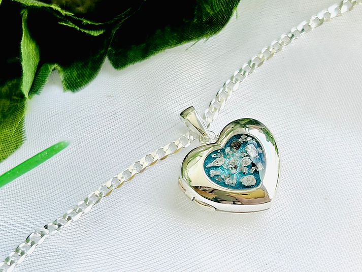 Silver heart-shaped pendant with blue and white floral inlay on a silver chain, resting on white fabric with green leaves nearby.