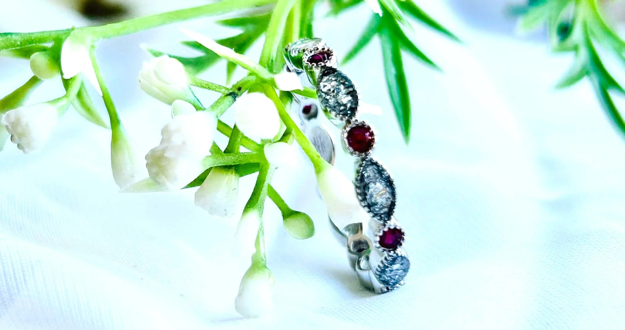 Close-up of a jewelry bracelet with dark red and silver stones resting on green leaves and white flowers.