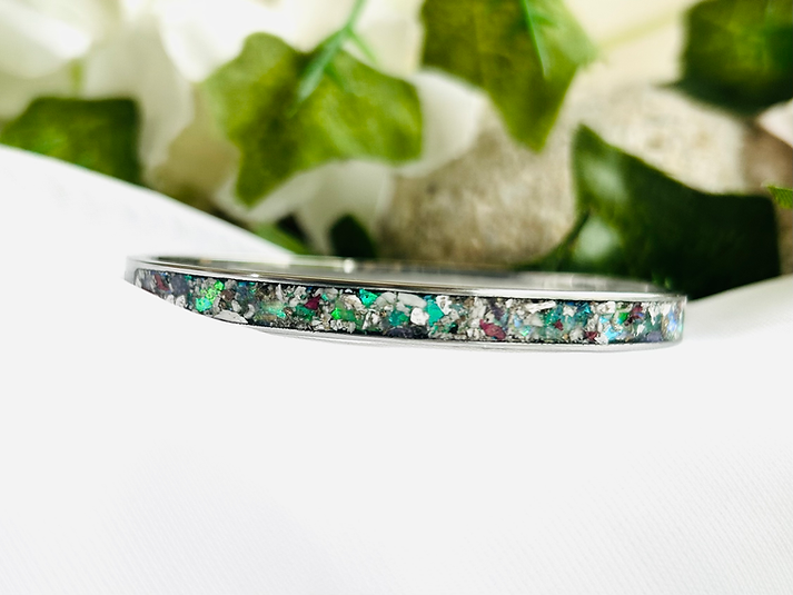 Close-up of a silver bracelet with colorful glittery stones, placed on a white surface with green leaves in the background.