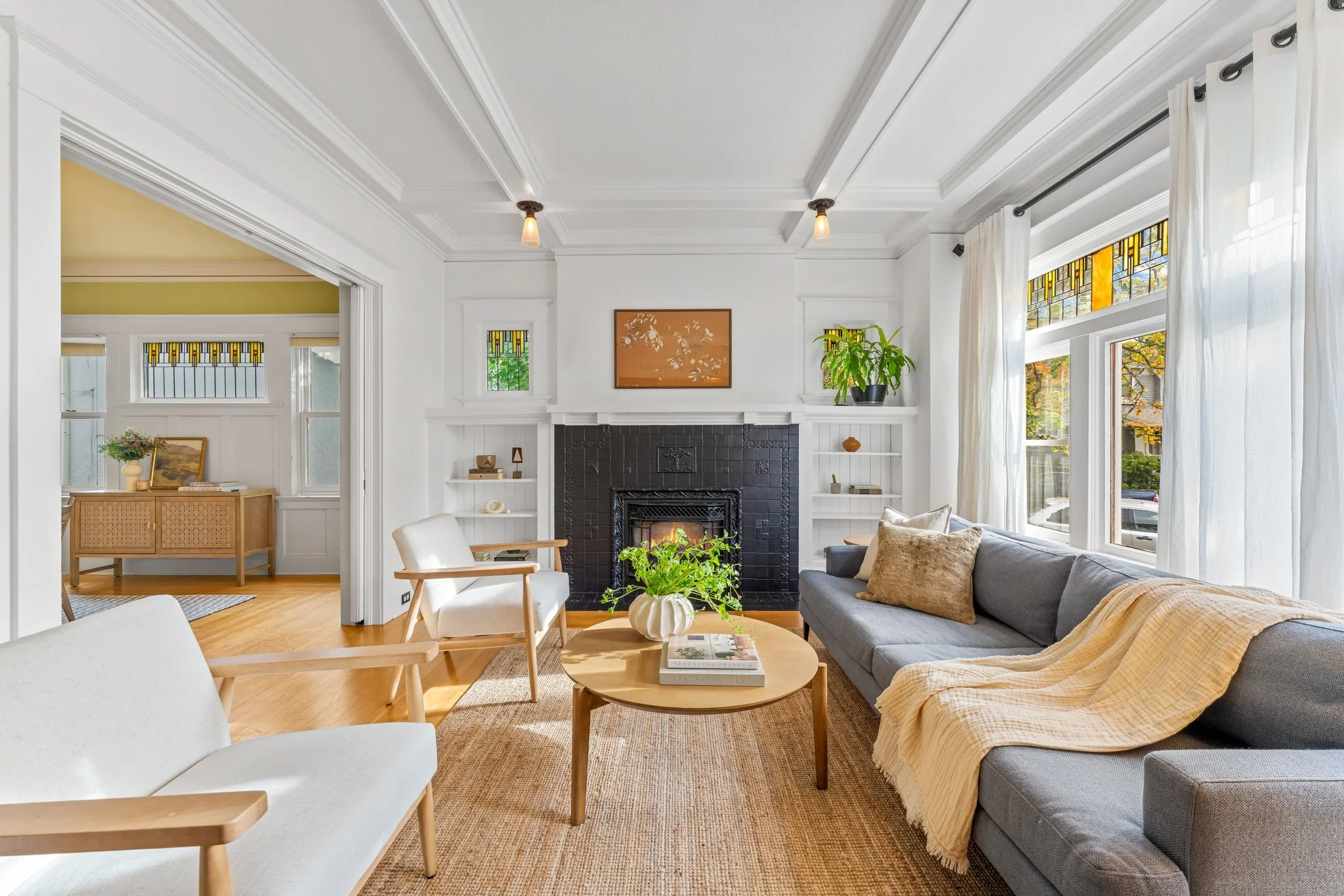 A living room with a white ceiling, white walls, large windows with white curtains, and a fireplace with a black tile surround. There are various pieces of furniture including a gray sofa with beige and tan pillows, white chairs, and a wooden coffee table with books and a plant. Decor includes a framed artwork above the fireplace and small decorative items on the built-in shelves.