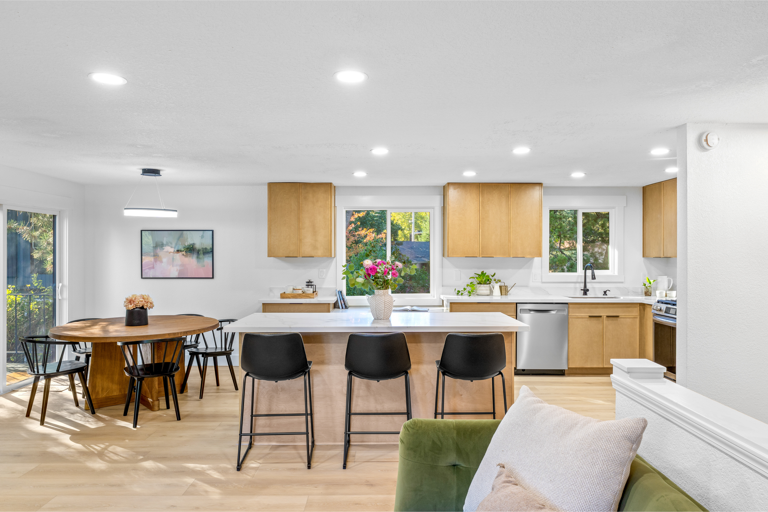 Modern kitchen and dining area with wooden cabinets, white countertops, and large windows showing outdoor greenery. There is a round wooden table with black chairs on the left and a kitchen island with black chairs in the center. A vase with pink flowers is on the table, and there are various kitchen items and plants on the counters.