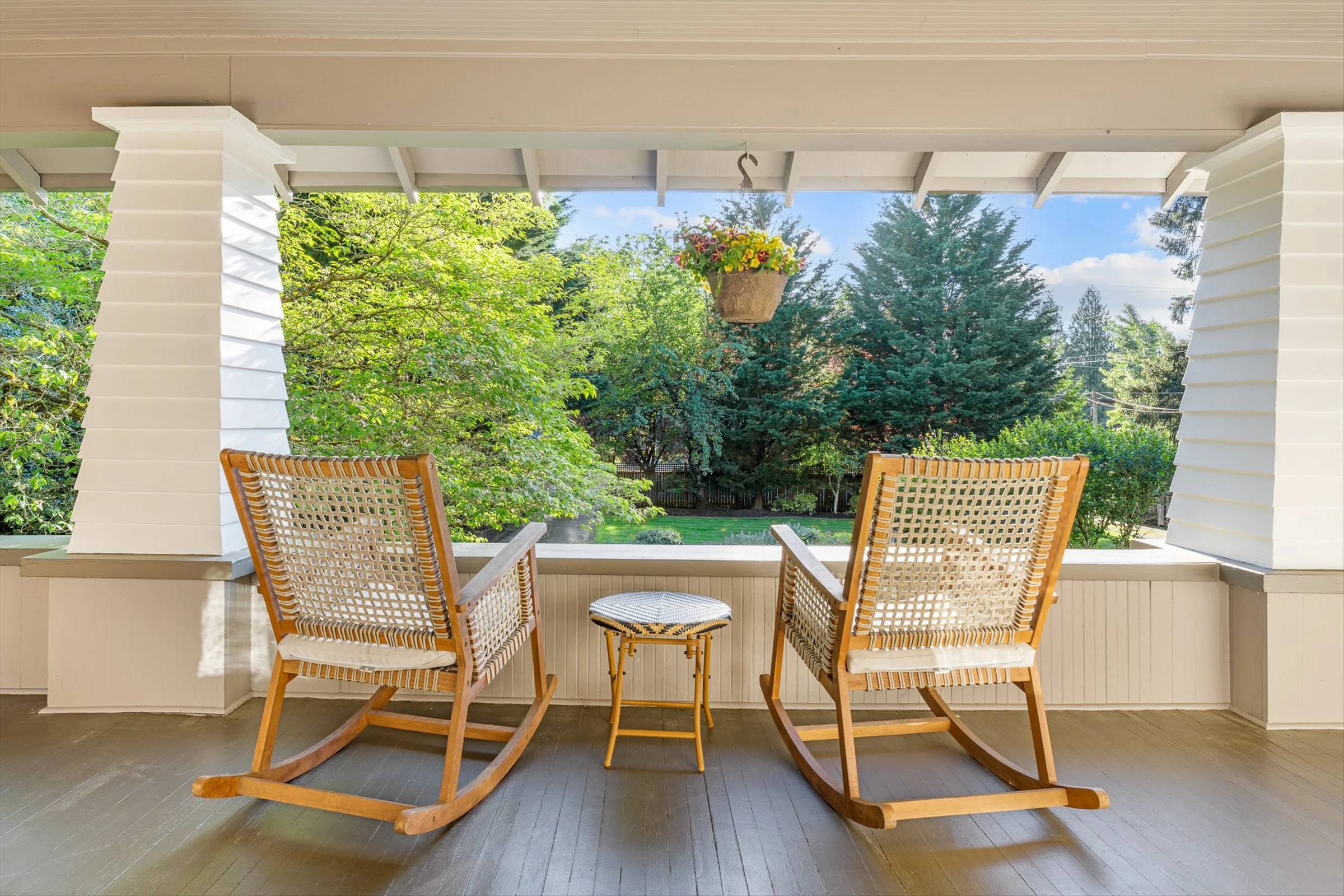 View of a porch with two wooden chairs, a small round table, and a hanging flower basket, overlooking a lush green backyard with trees and a blue sky.