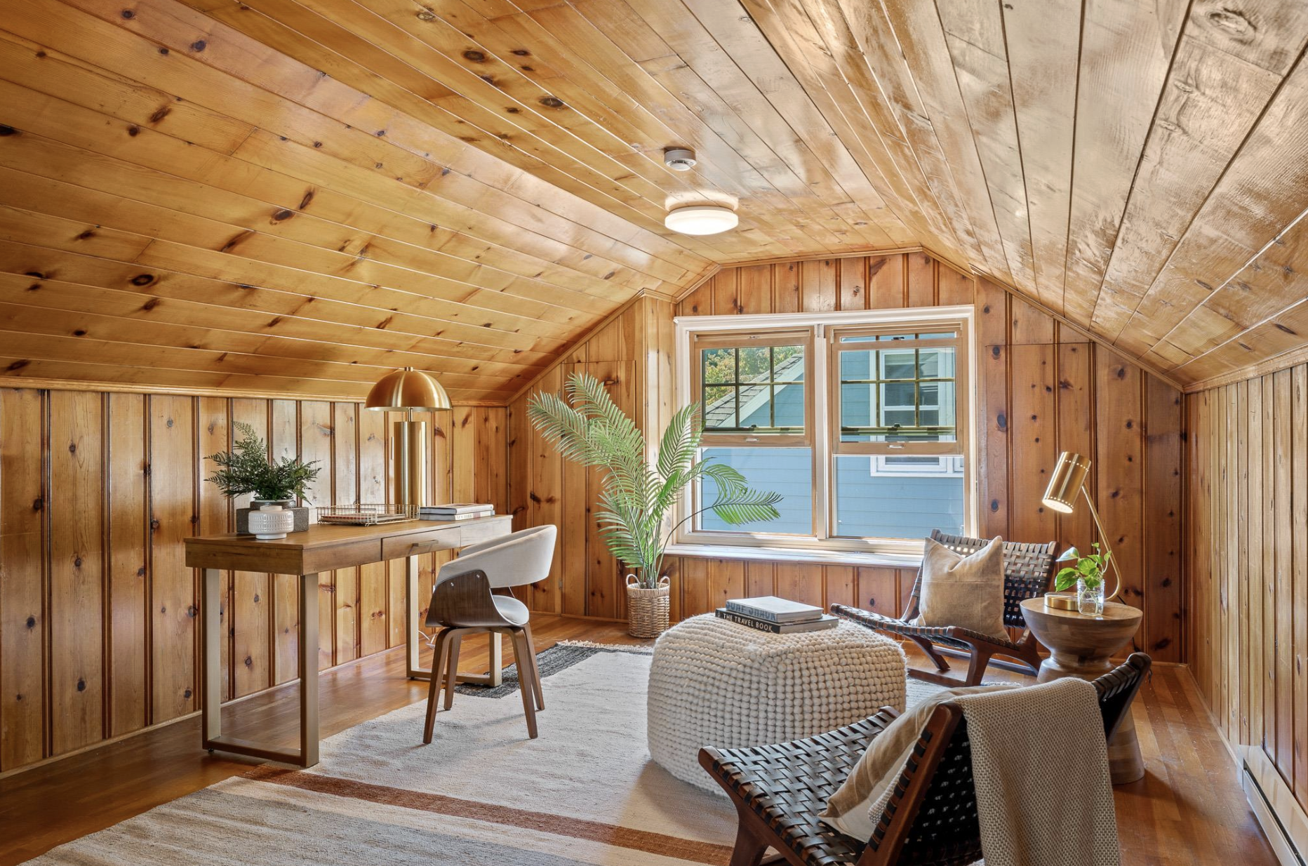 A cozy room with wood-paneled walls and ceiling, featuring a window, houseplants, a cozy chair, a small table, and modern lighting fixtures.