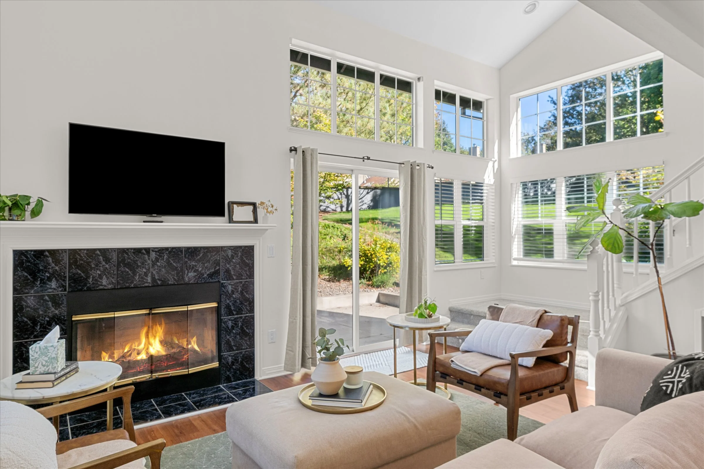 Bright living room with large windows, white walls, and modern decor, featuring a fireplace, plants, and seating area.