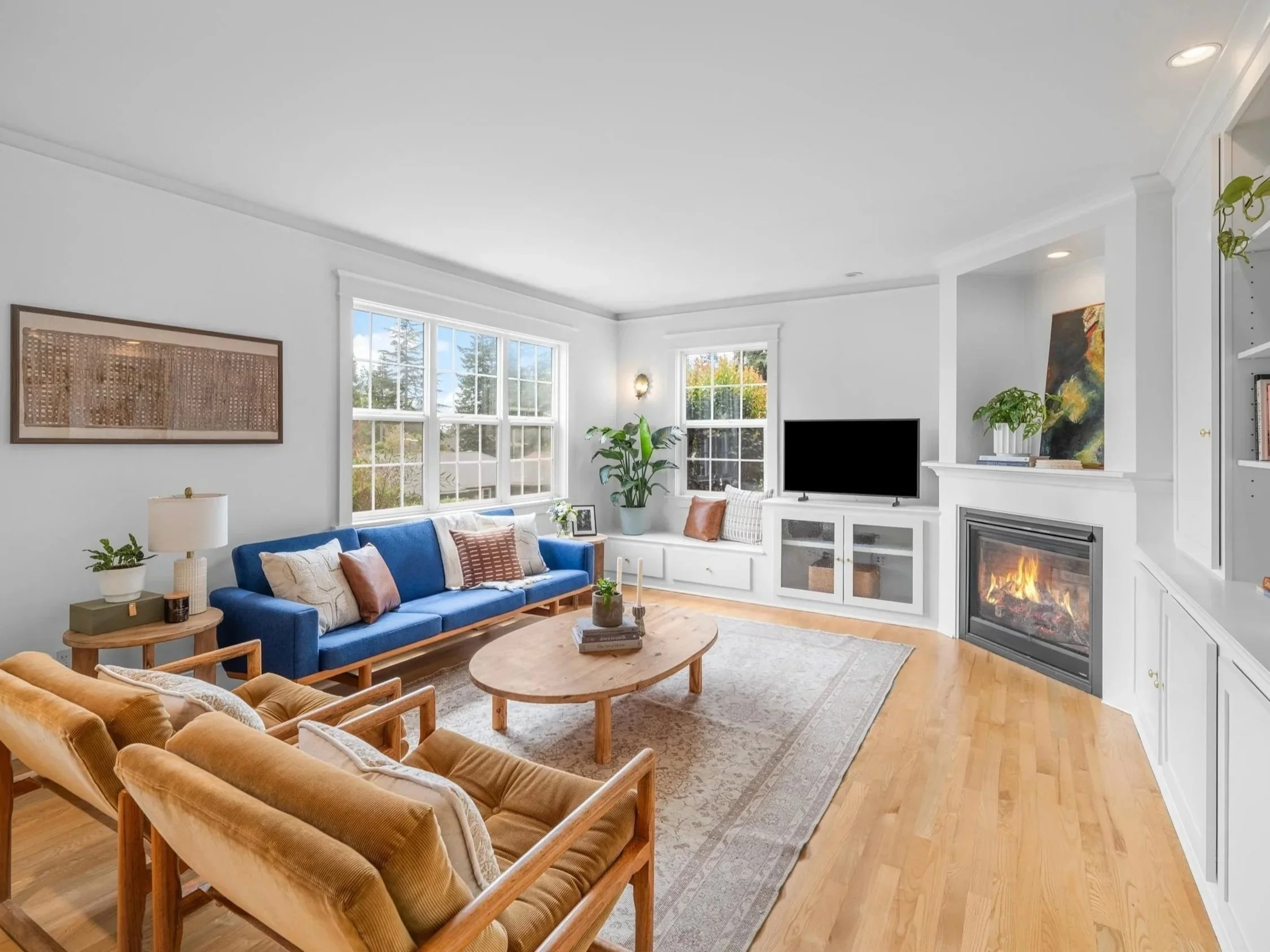 A bright living room with white walls and large windows showing trees outside. It features a blue sofa with beige and brown pillows, and a wooden coffee table. There's a fireplace with a painting above it, various plants, and a beige carpet on a light wood floor.