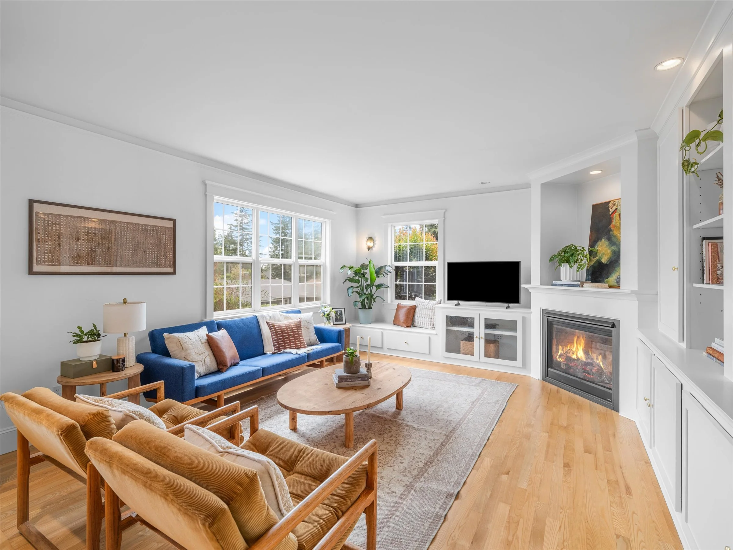 Living room with white walls, large windows, a blue sofa, tan armchairs, a wooden coffee table, a fireplace, a TV, and various plants and artwork.