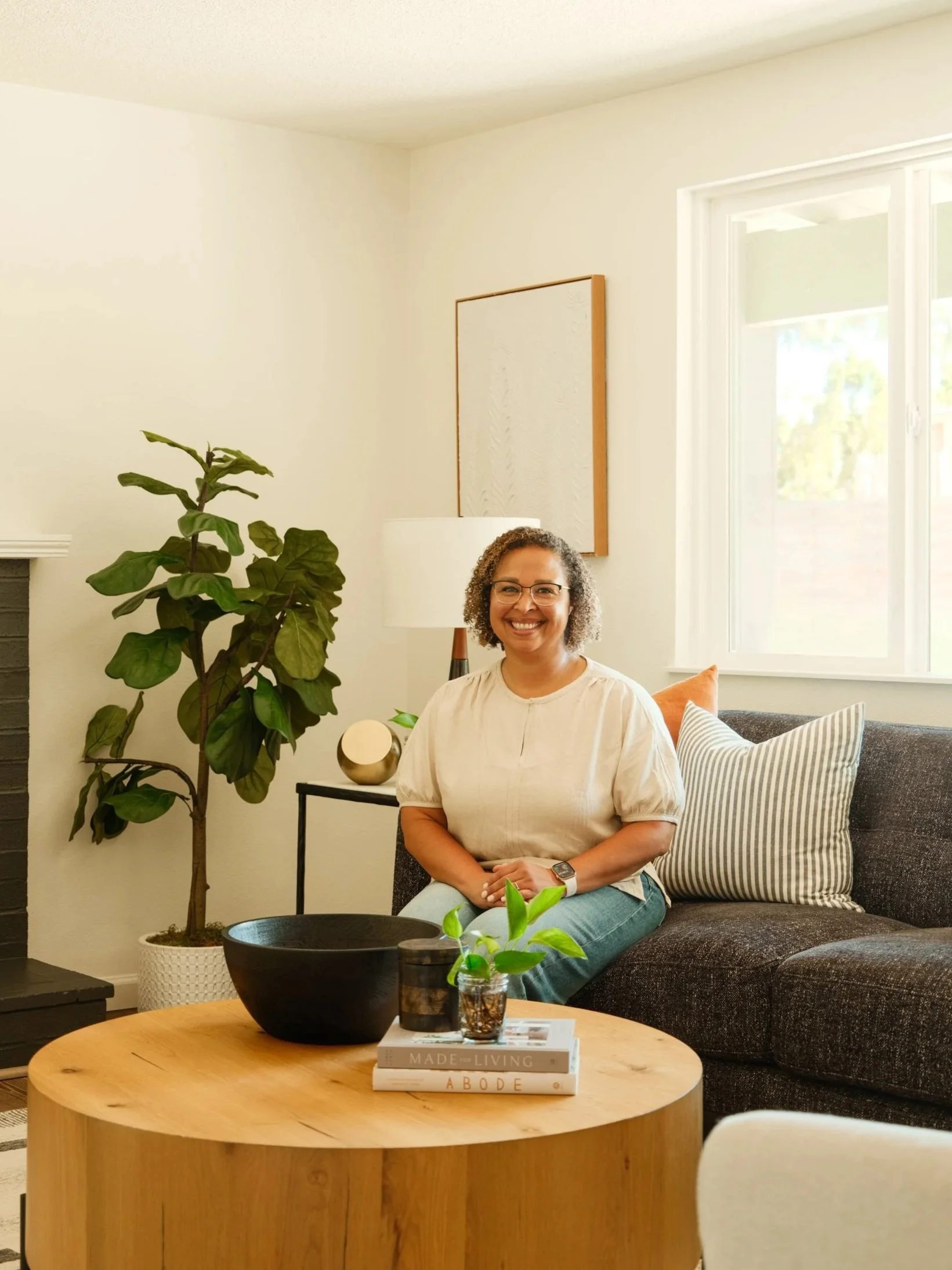 A woman with curly hair and glasses sitting on a black couch in a living room, smiling at the camera. The room has large windows, a potted plant, a floor lamp, and a side table with decorative items and books.