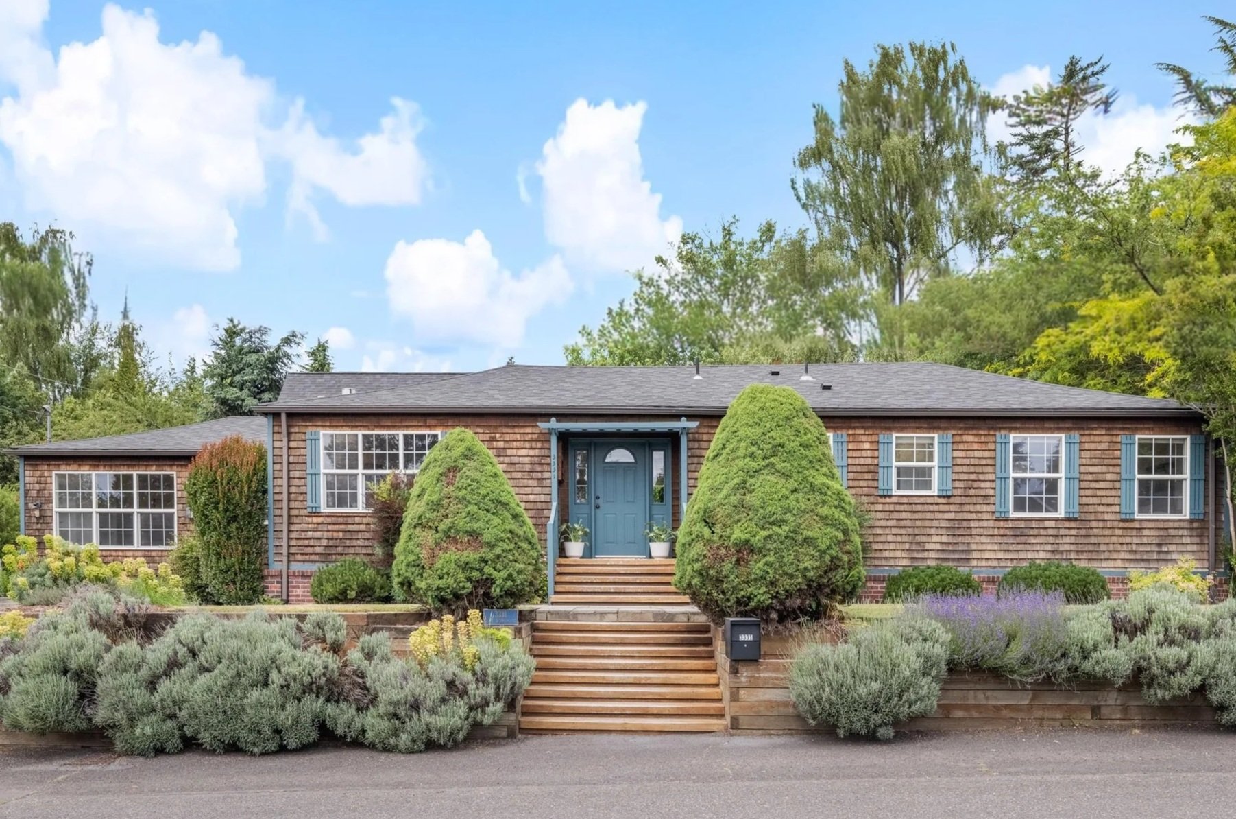 A single-story house with a gray roof, wood siding, blue shutters, a blue front door, and a front porch with steps. The house is surrounded by landscaped bushes and plants, with a garden bed and trees in the background under a blue sky with clouds.