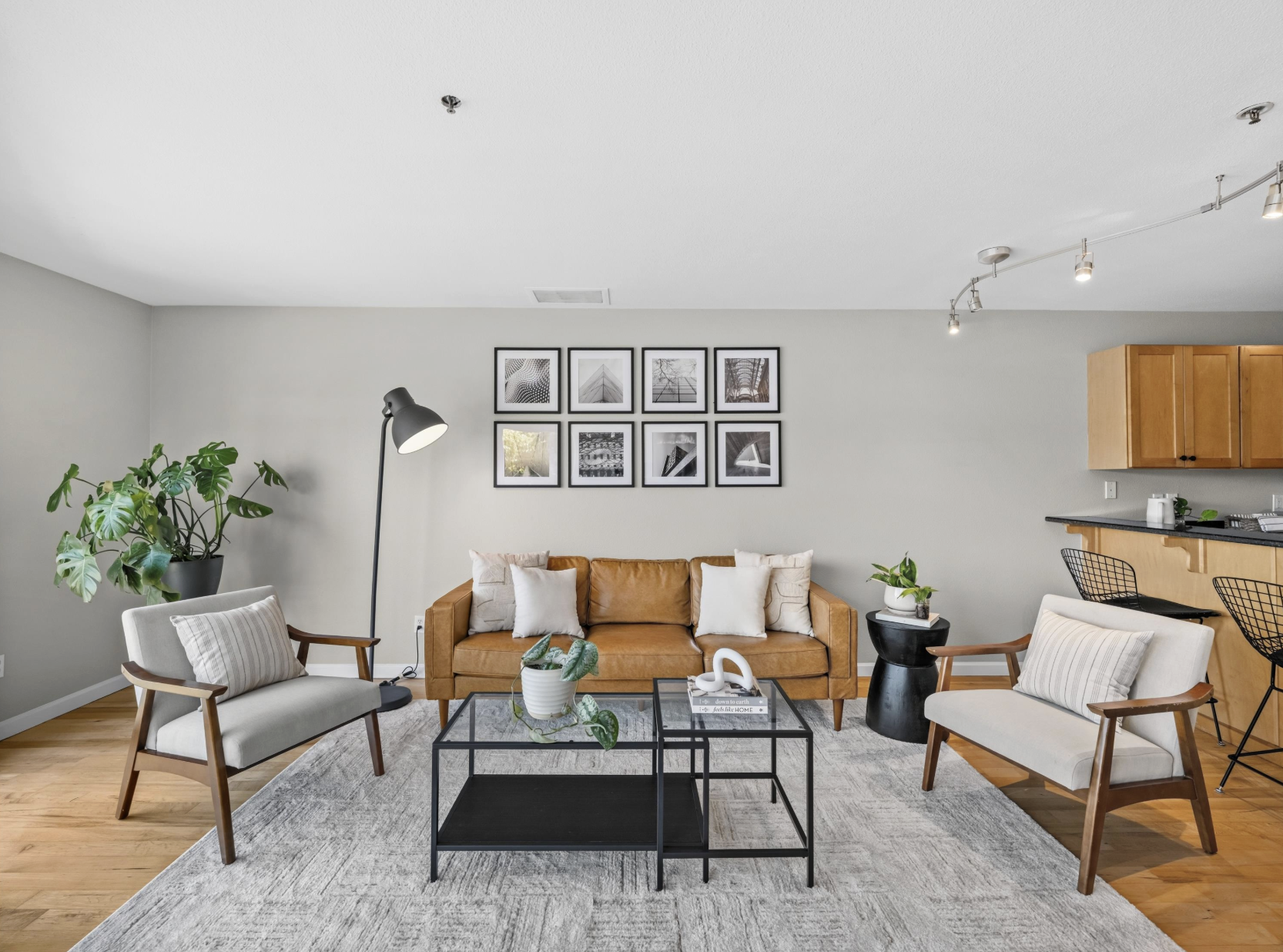 A modern living room with a brown leather sofa, white pillows, and a black coffee table on a light gray rug. Two white armchairs with wooden frames sit nearby, with a large green plant to the left. A black floor lamp is beside the sofa. The wall above the sofa has nine black-and-white framed photographs arranged in three rows. To the right, part of a kitchen with wooden cabinets and a black countertop is visible, with a bar area and black wire chairs.