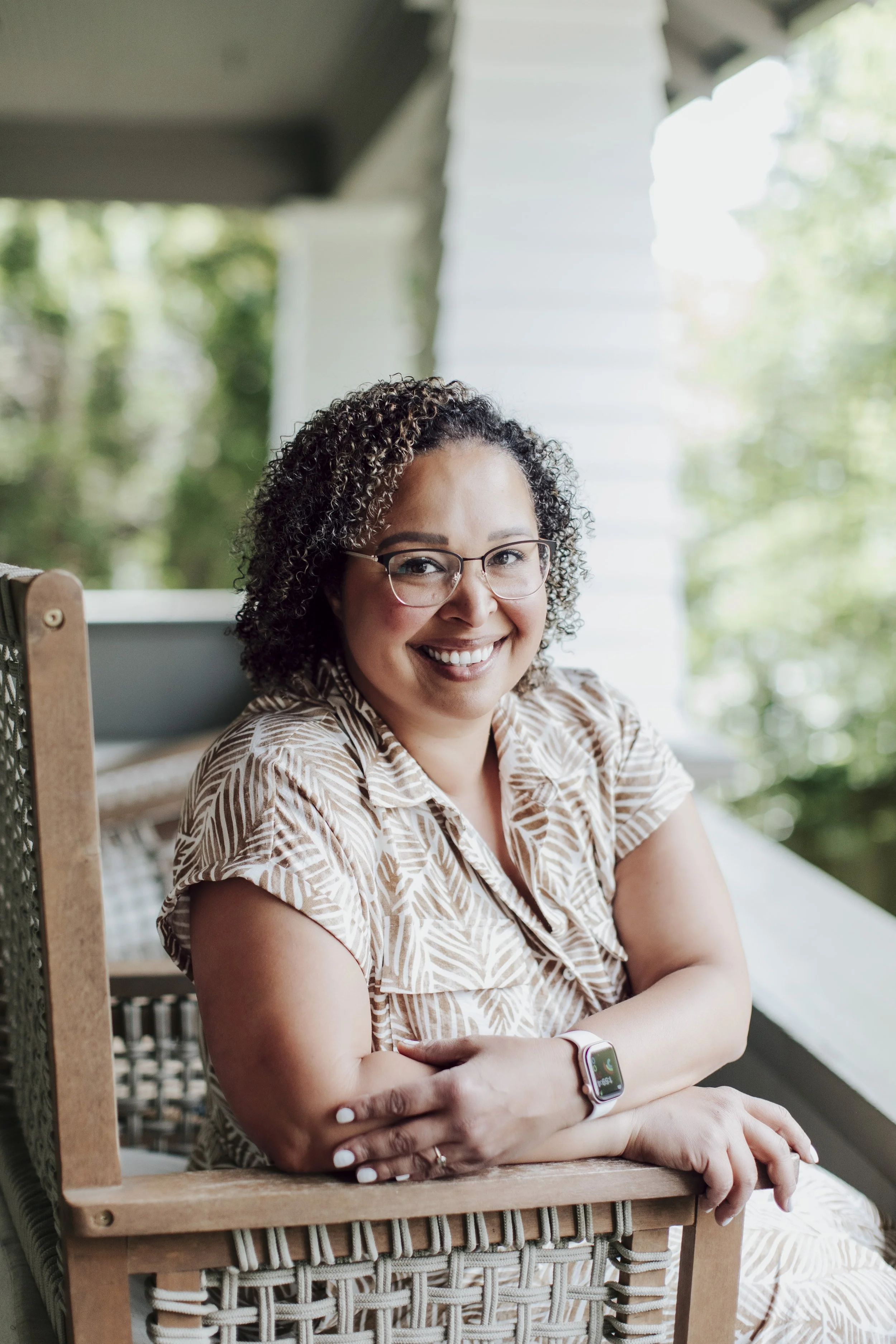 A woman with curly hair, glasses, and a smartwatch, smiling and sitting with crossed arms at a rustic outdoor setting.