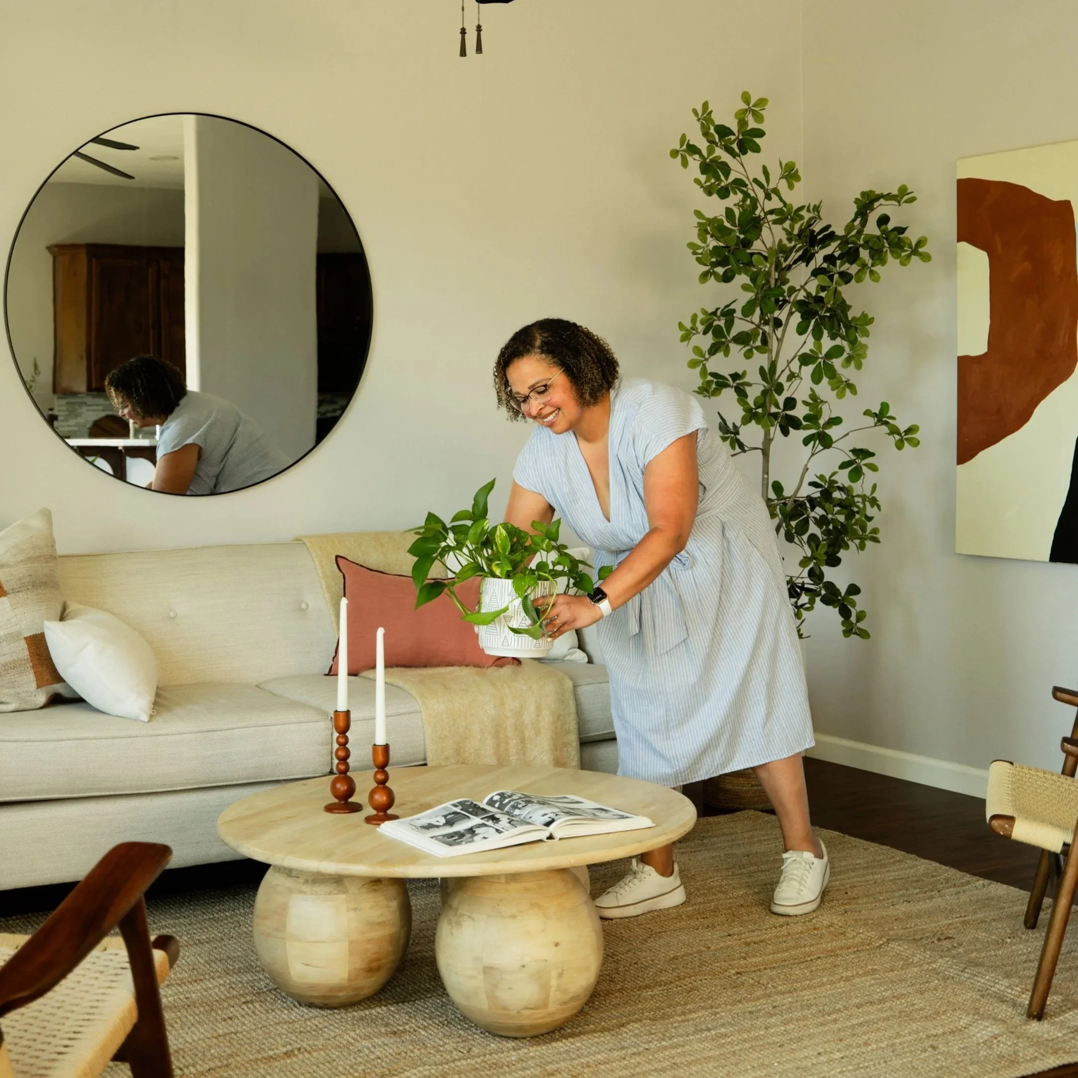 A woman with curly hair and glasses is arranging a plant in a white decorative pot on a beige living room table. A large round mirror reflects her back, showing a wooden cabinet in the background. The room has a light-colored wall decorated with a large abstract painting and a green indoor plant.