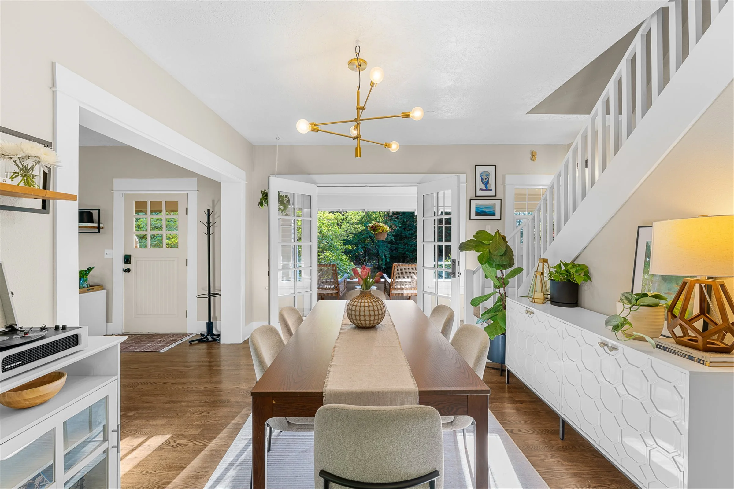 Bright dining room with open back doors leading to a patio surrounded by green trees, a modern chandelier hanging from the ceiling, decorative plants and framed artwork on the walls, and a light wood dining table with chairs.