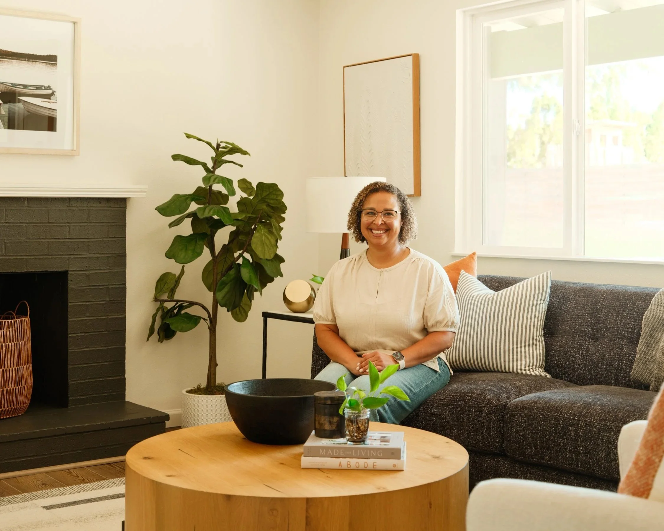 A woman with curly hair and glasses smiling while sitting on a dark-colored couch in a living room with white walls, large windows, a wooden coffee table with books and plants, and a large plant beside her.