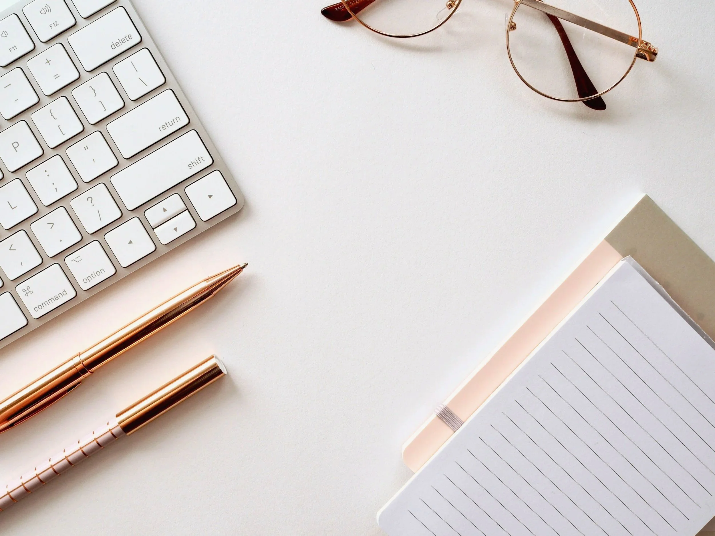 A white keyboard, a pair of gold-rimmed eyeglasses, two rose gold pens, a notebook with lined pages, and a closed book on a white surface.