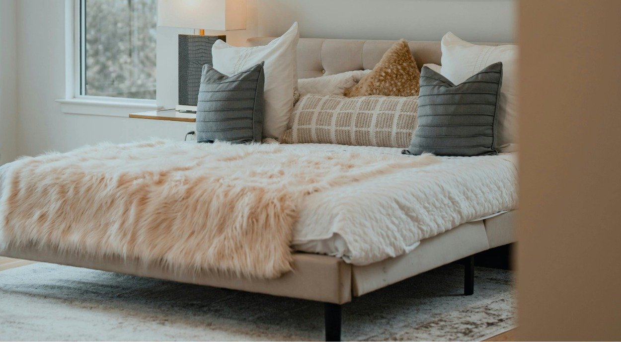 A cozy bed with white bedding, a light tan faux fur blanket, and multiple pillows in shades of white, beige, and gray.