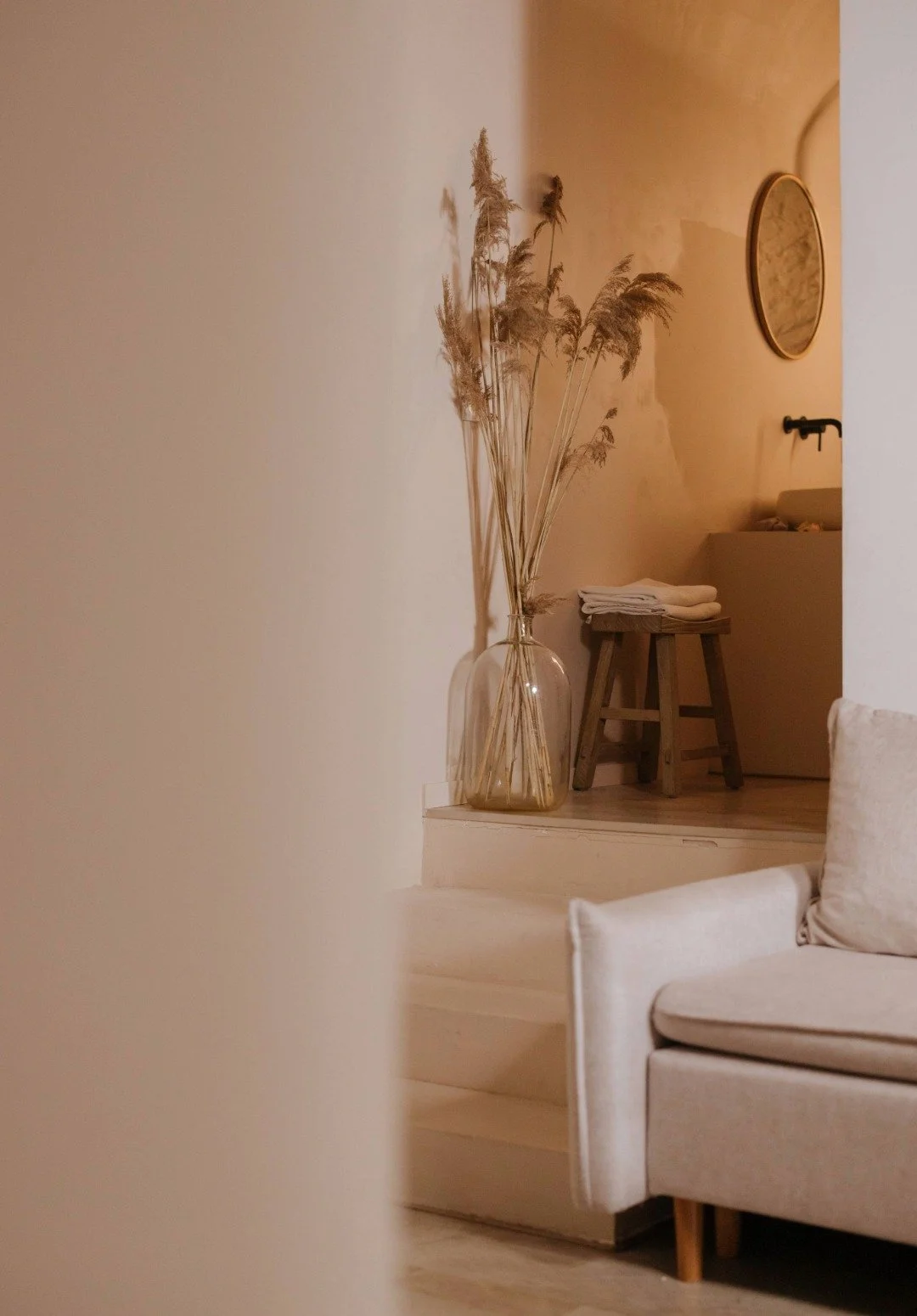 A cozy interior with a beige sofa, a wooden stool with folded towels, a large glass vase with dried pampas grass, and warm lighting.