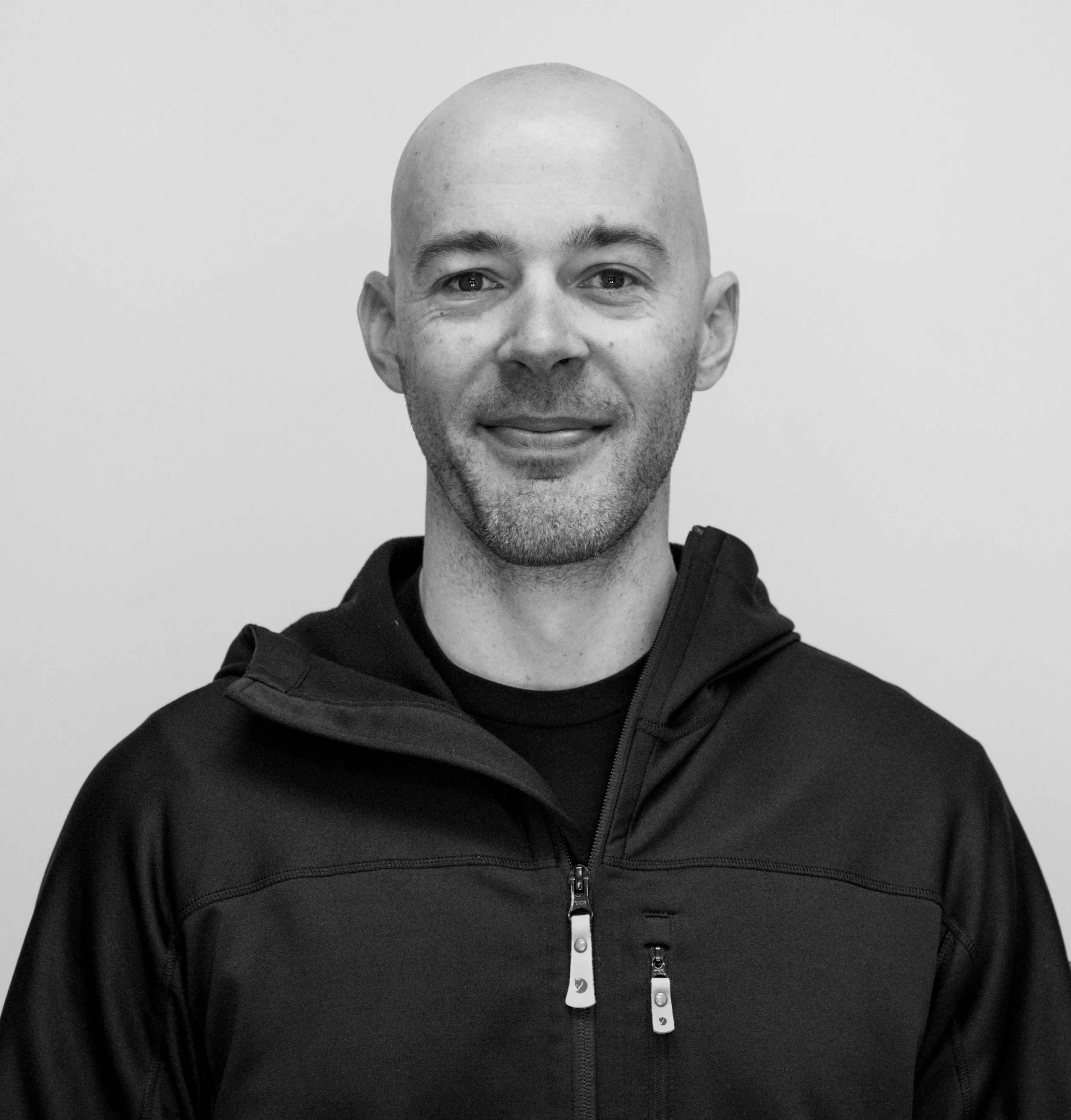 Black and white portrait of Andrew Parrott, smiling, wearing a black zip-up jacket with zippers.