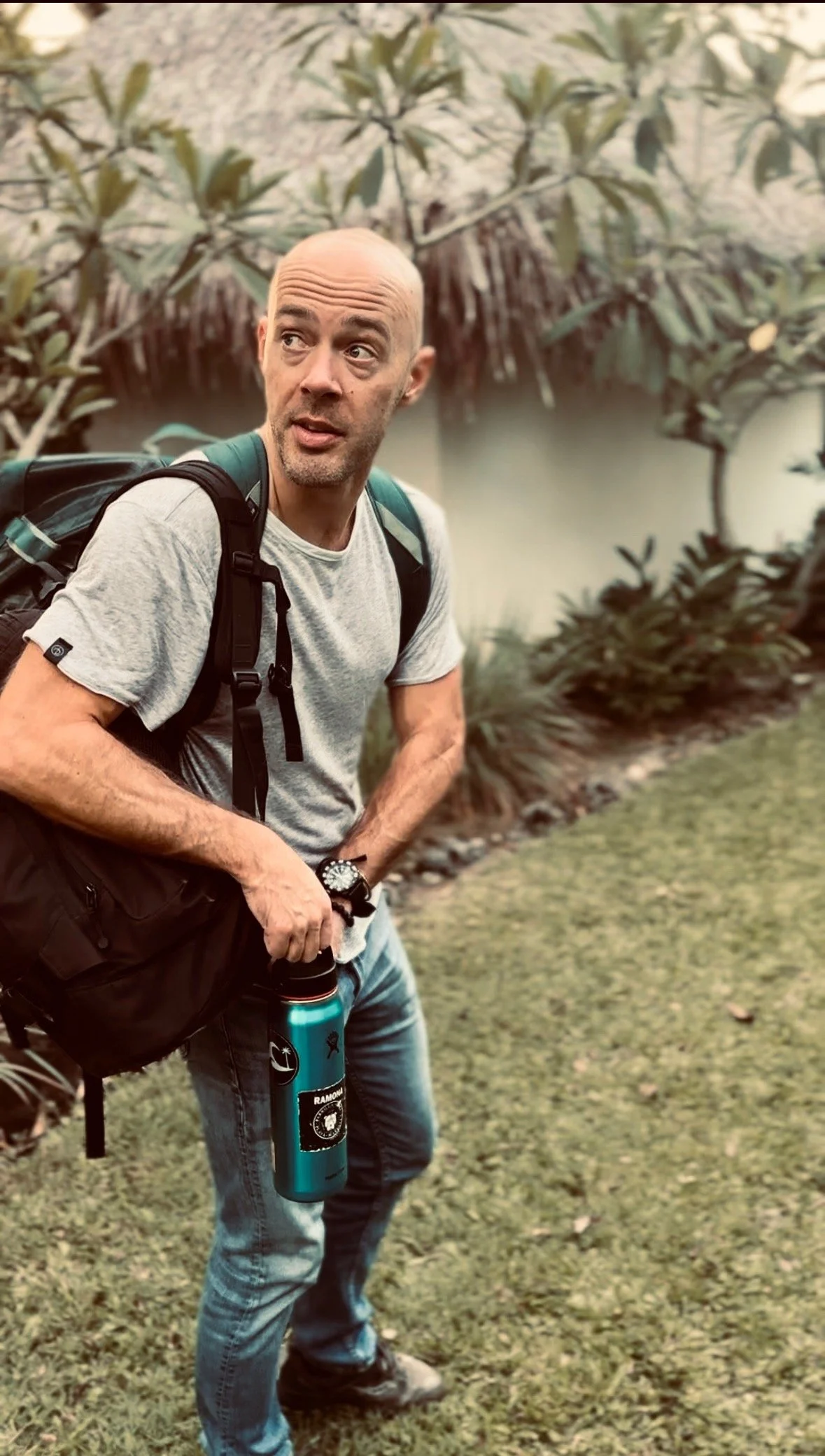 Andrew Parrott with a beard and casual clothes holding a blue water bottle, carrying a backpack, standing on a grassy area with plants and trees in the background.