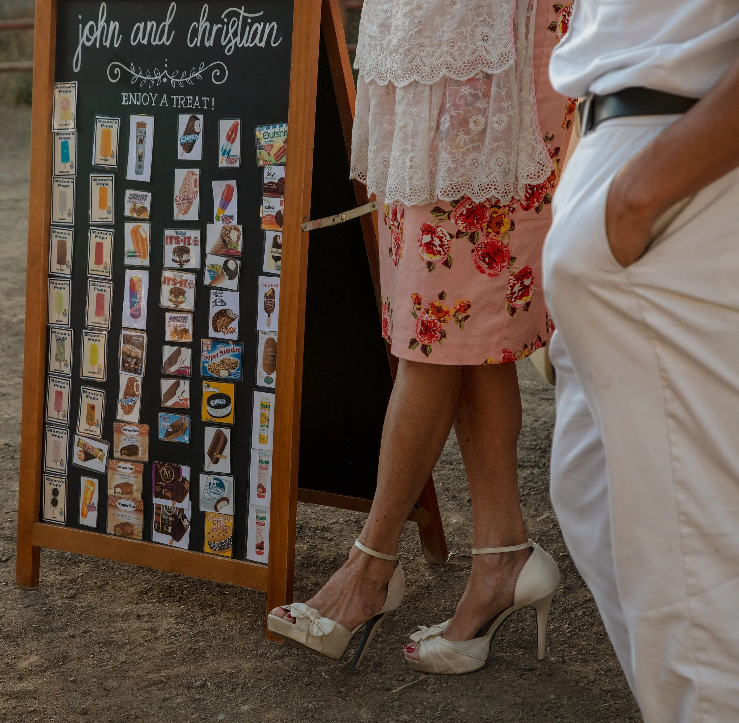 A person in white shoes and cream-colored shorts stands next to a woman wearing pink floral shorts and a white lace top, next to an outdoor menu display with images of various ice cream treats.