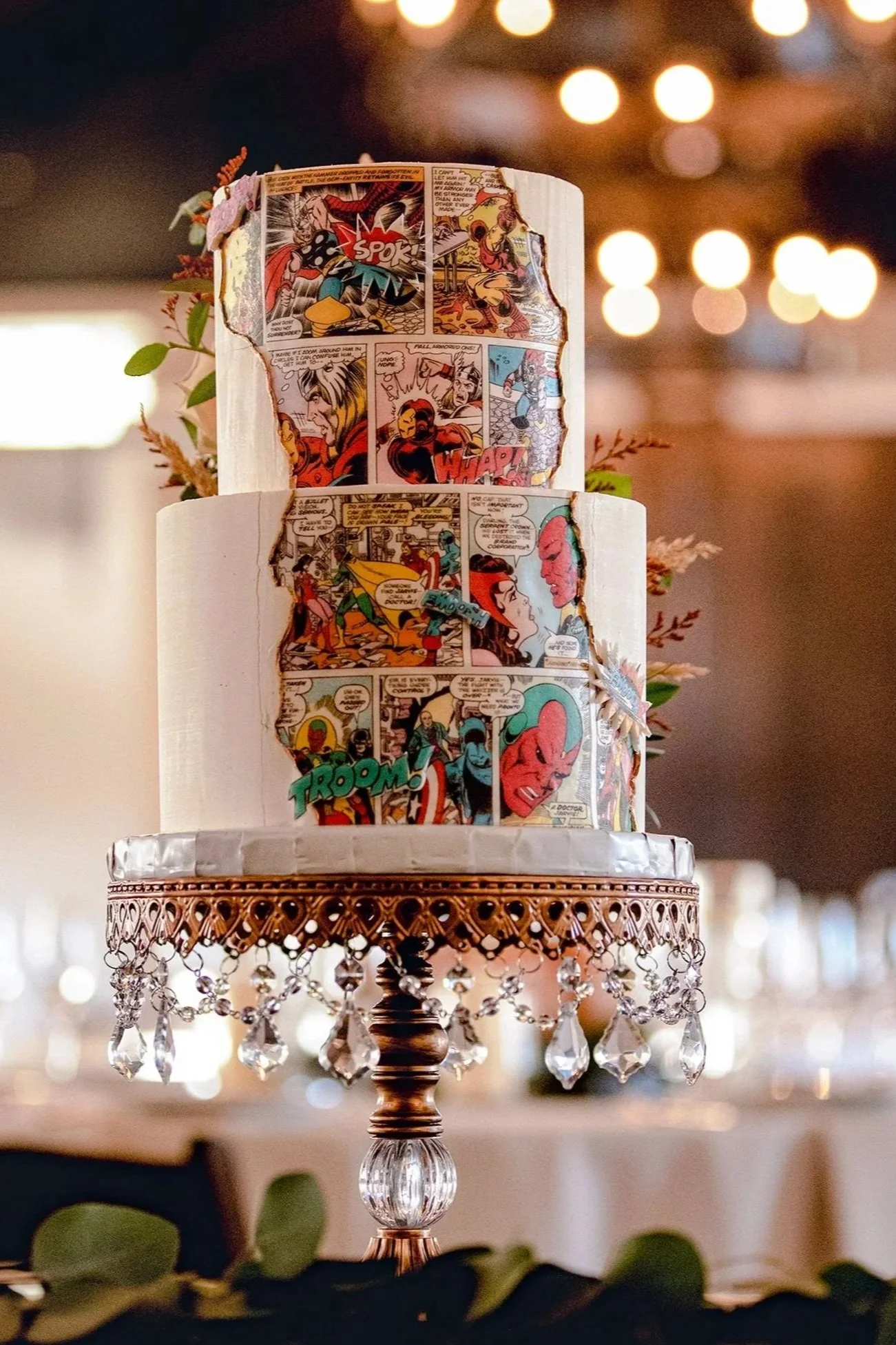 A decorative cake is decorated with Batman comic strip images and has a lace and crystal beaded trim at the base.