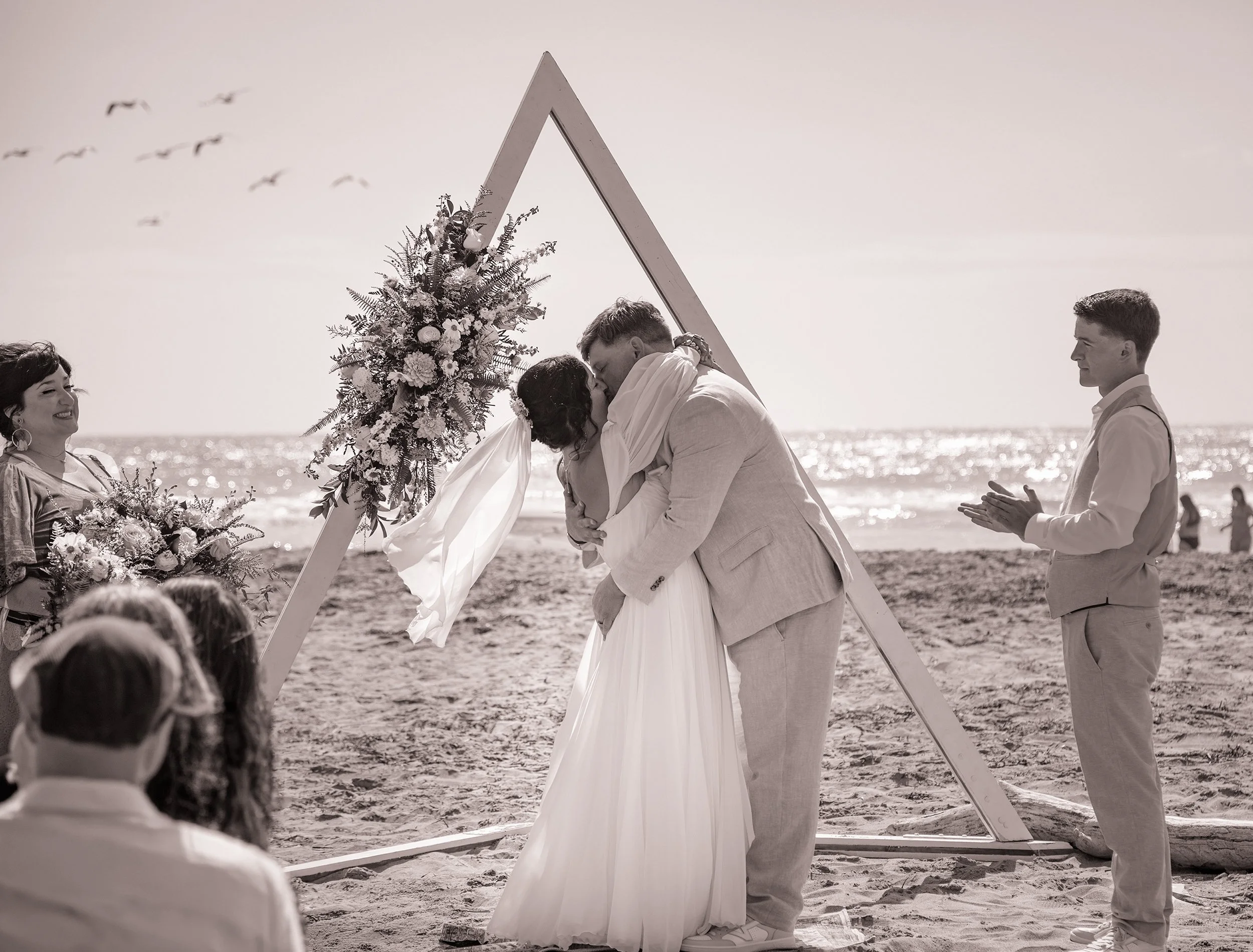 A couple sharing a kiss during a beach wedding ceremony, with an officiant and guests, overlooking the ocean.