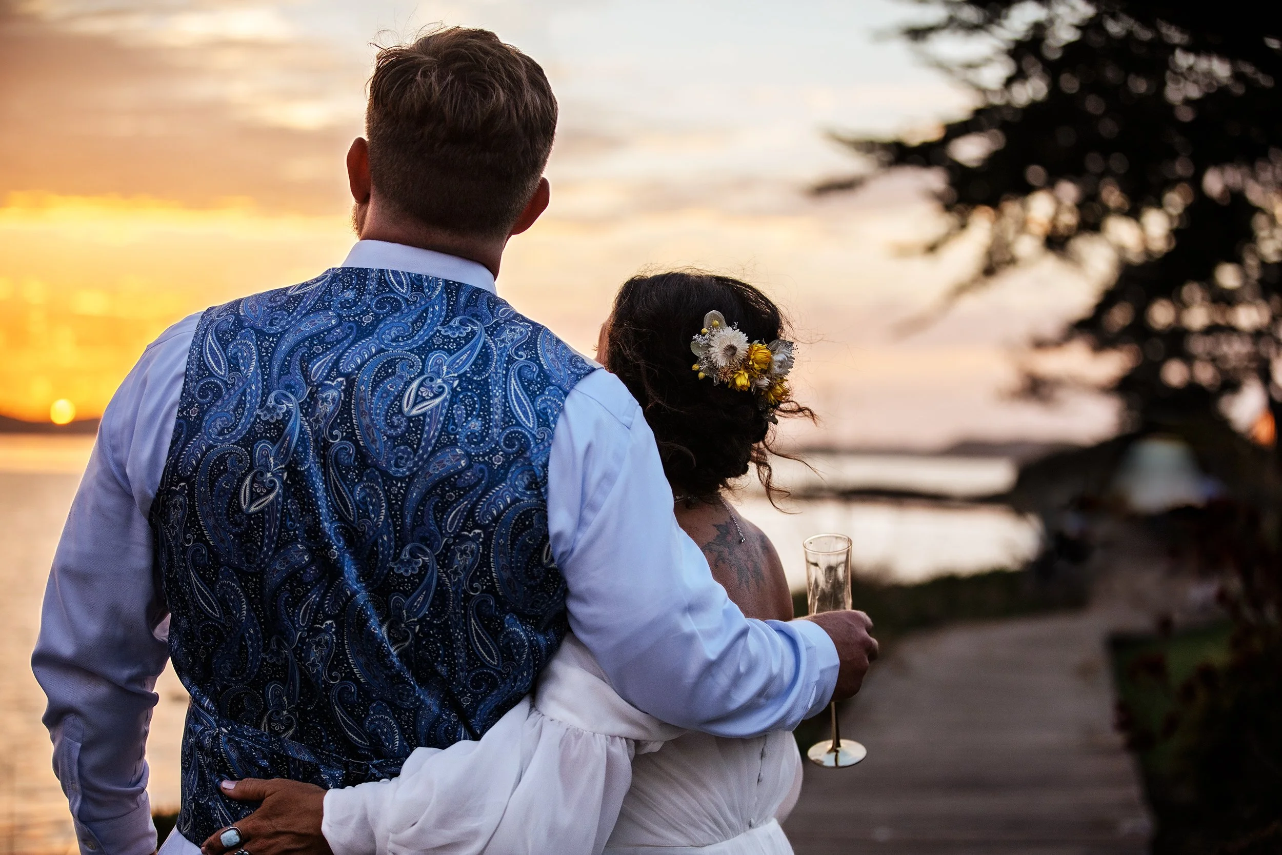 A couple dressed in wedding attire, standing together outdoors at sunset, with the man’s arm around the woman as they look at the sunset over a body of water.