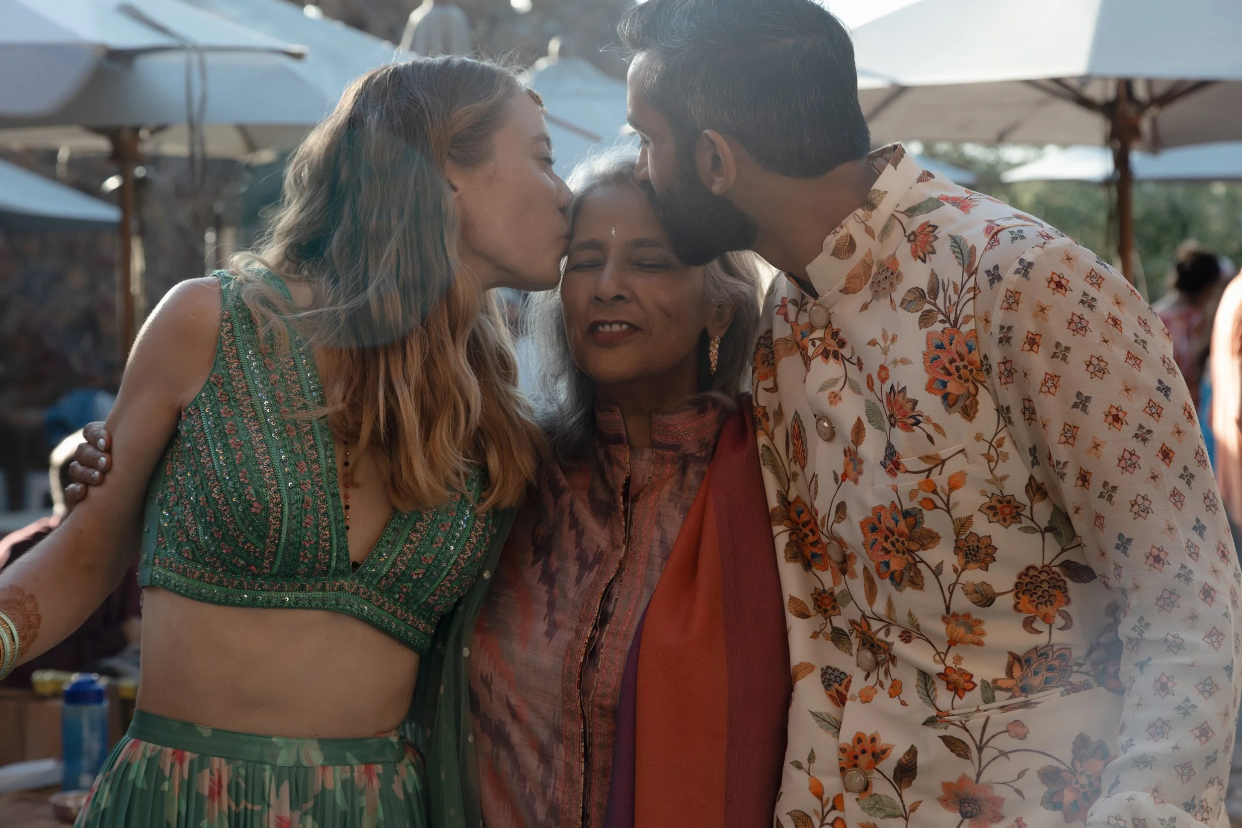Three people, two women and one man, are close together, with eyes closed and foreheads touching, in a warm, outdoor setting with umbrellas and people in the background.