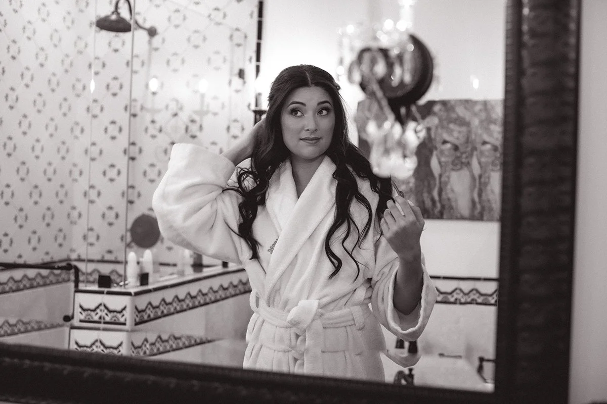 A woman with long dark curly hair taking a selfie in a mirror. She is wearing a white bathrobe and standing in a decorated bathroom with patterned tiles and artwork.