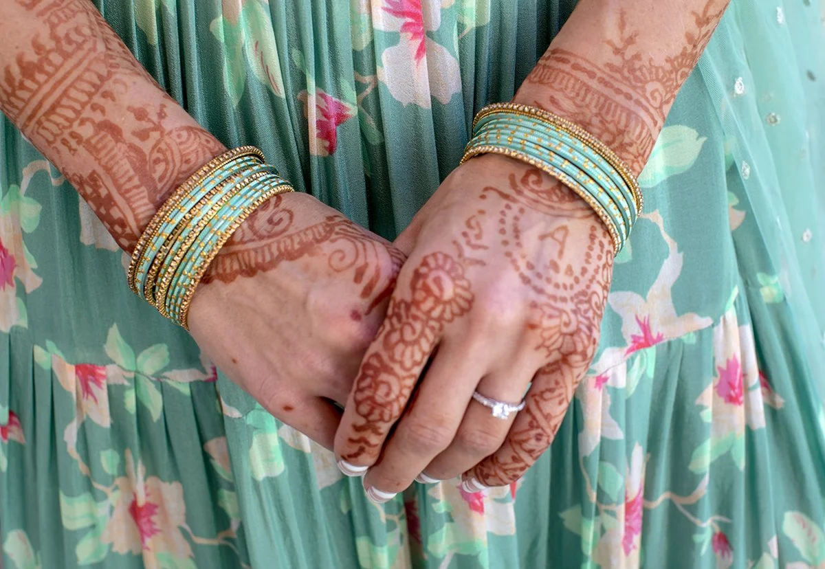 Close-up of a woman’s hands and forearms adorned with colorful bangles and a ring, displaying intricate henna tattoos, with a floral and green fringed dress in the background.