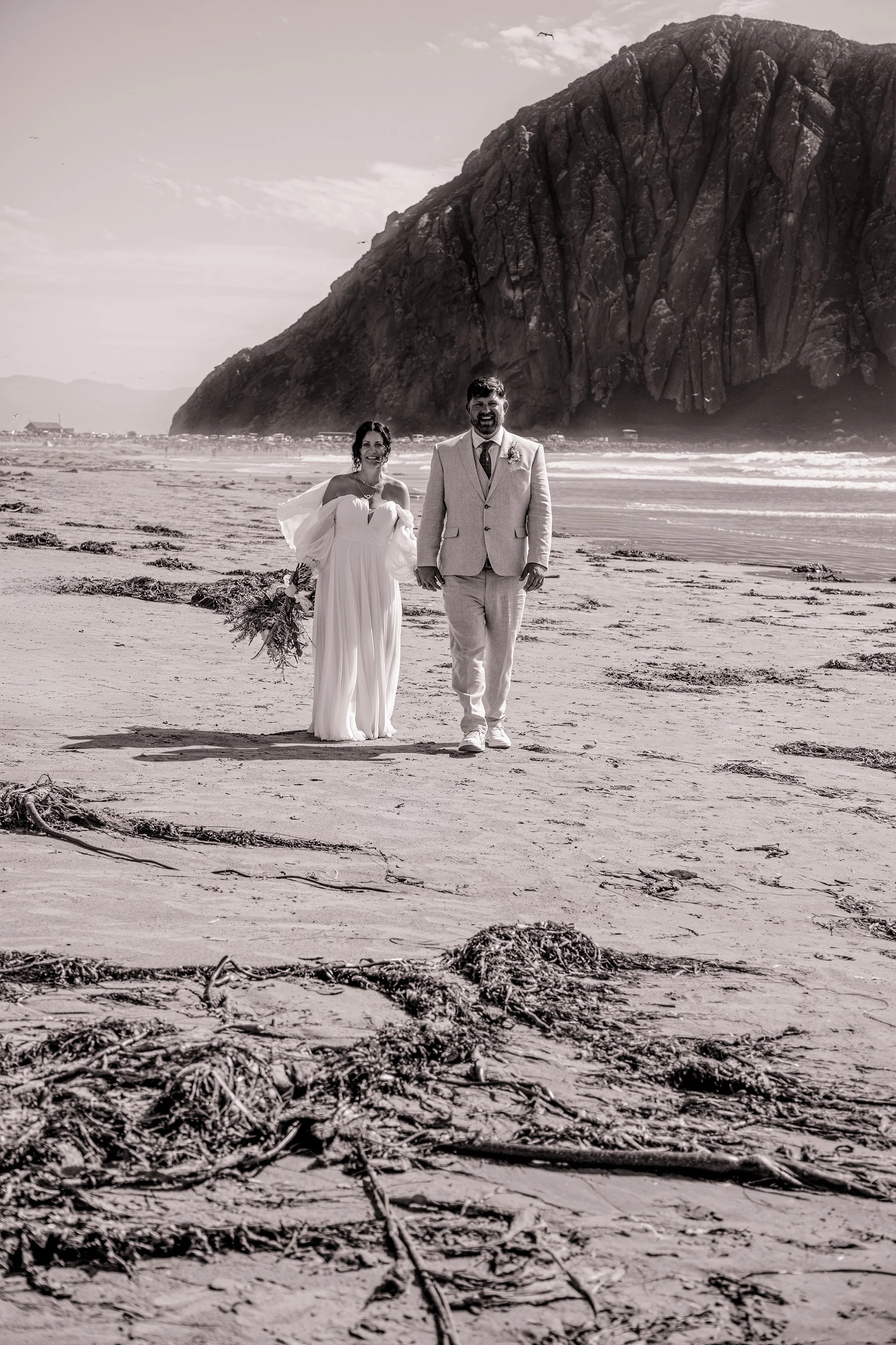 A couple in wedding attire walking on the beach near a large rocky cliff, with a clear sky and a bird flying overhead.
