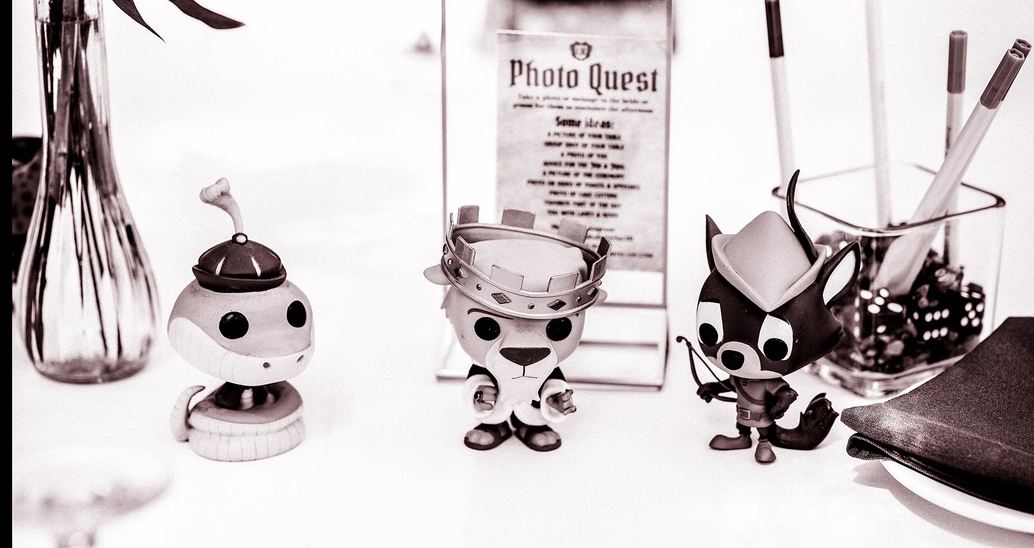 Collection of small toy animals and objects on a table, including a cat, a dog, and a panda with various accessories, near a basket of pens and a framed sign.