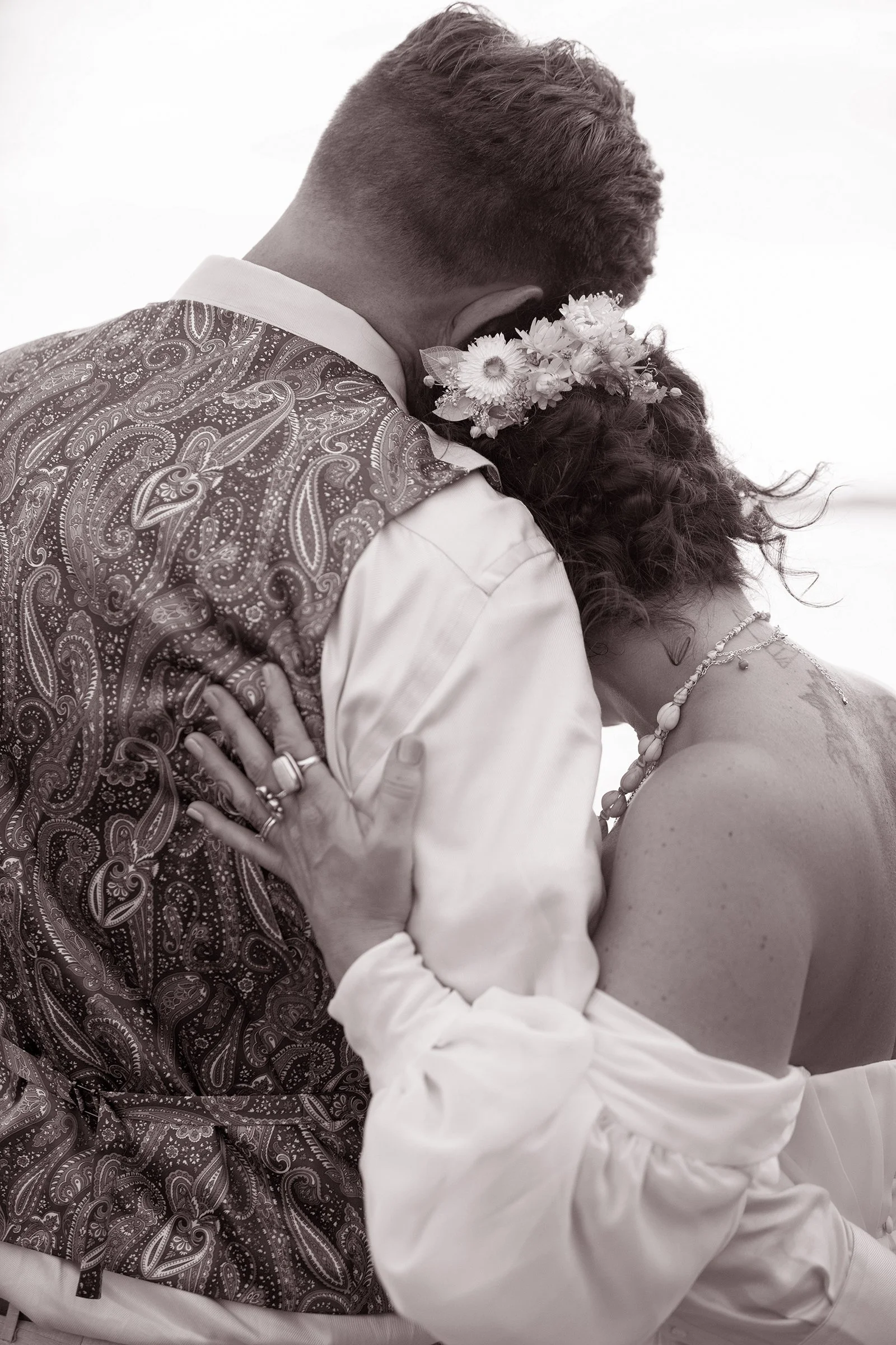 A man and woman hugging closely, the woman wearing a flower crown and pearl jewelry, the man dressed in a patterned vest and white shirt.