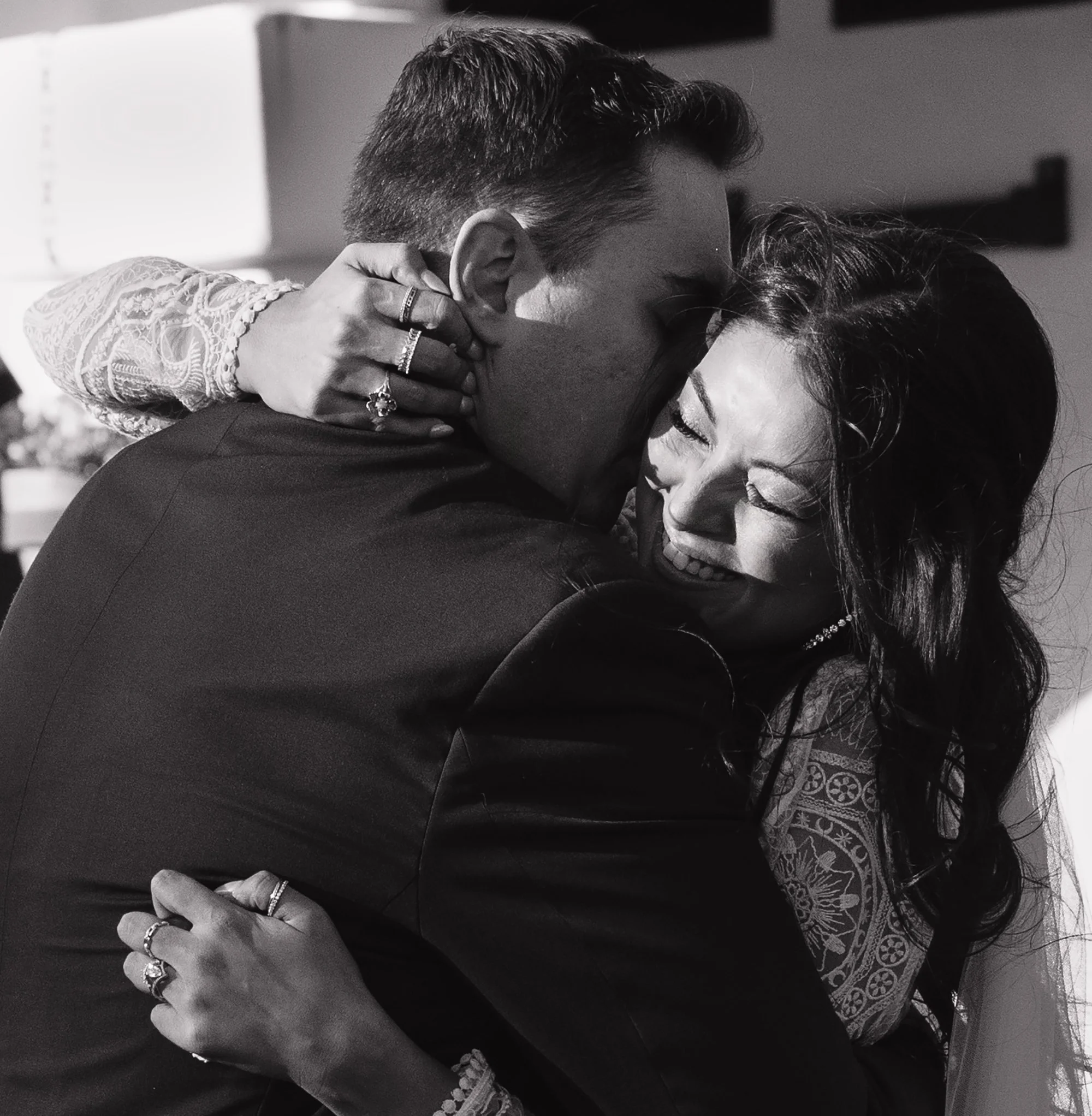 A black and white photo showing a couple in an intimate pose, with the man holding the woman close and their faces touching. The woman is wearing decorative earrings and rings, and a patterned dress or shawl. The photo captures a moment of affection.