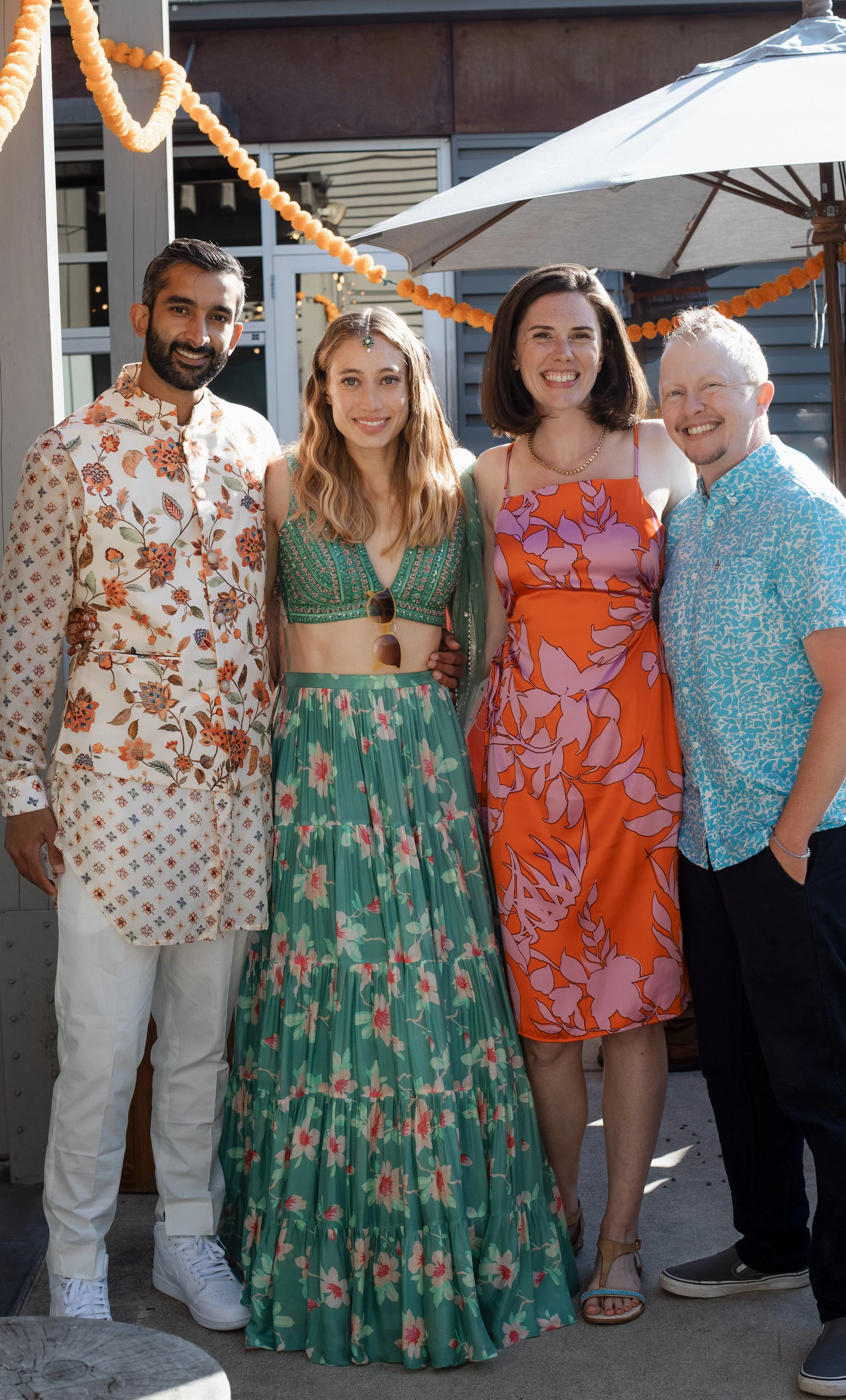 Group of four people smiling at a social gathering outside, dressed in colorful summer clothing, with decorations and a patio umbrella in the background.