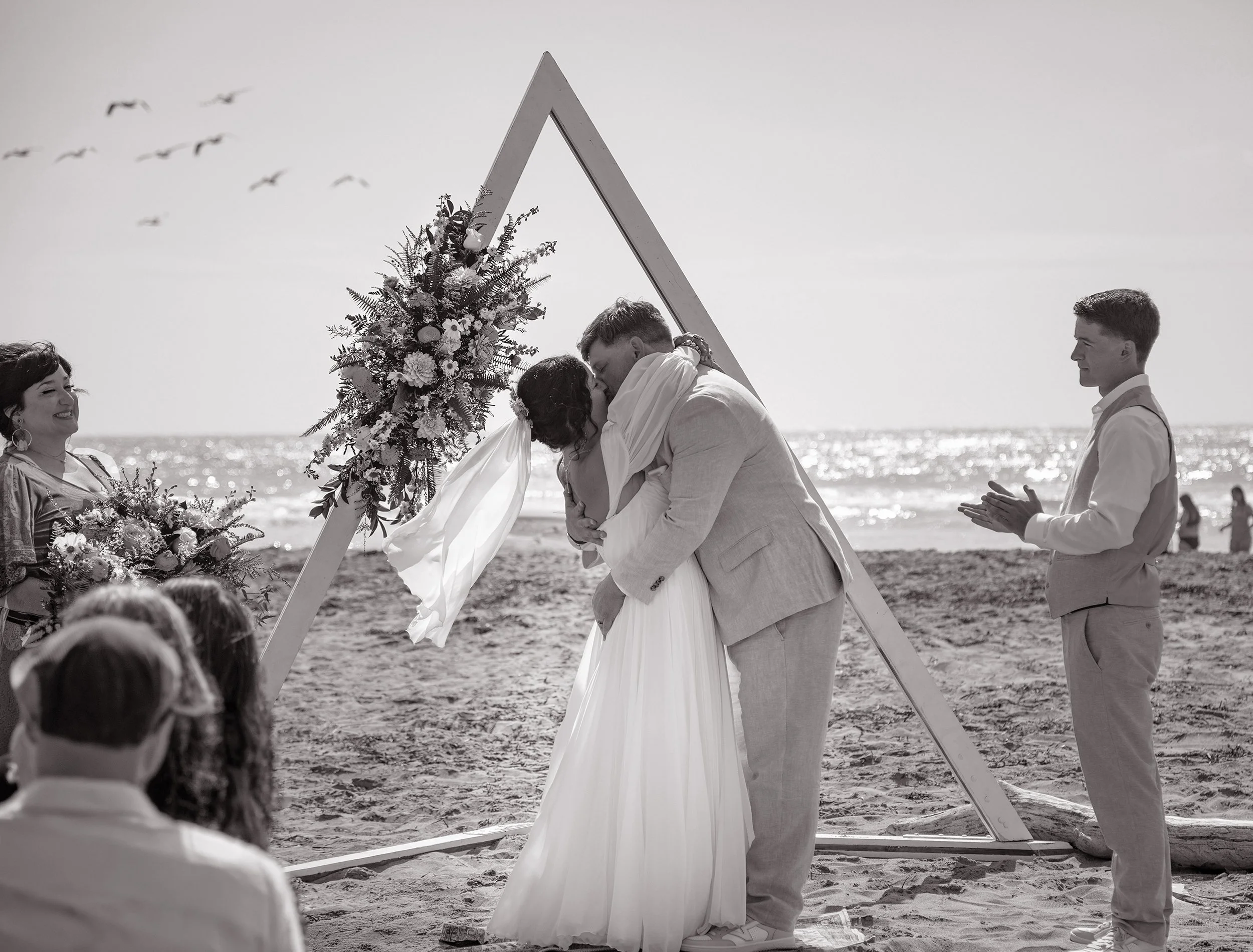 A couple exchanges a kiss at a beach wedding ceremony, with an officiant and a young man clapping nearby. There is a wooden arch with flowers behind them and seagulls flying in the sky.