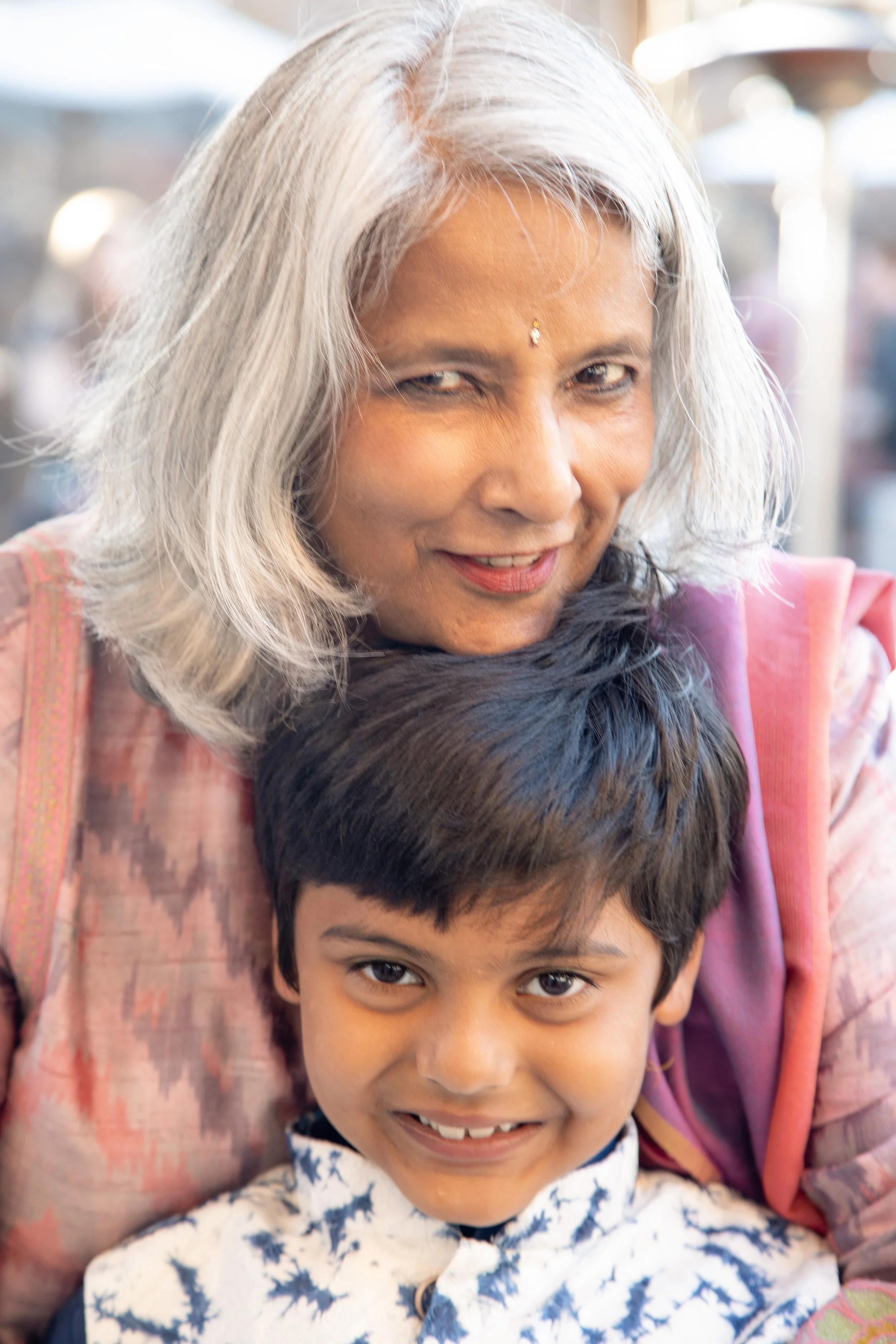 A smiling elderly woman with long gray hair and a bindi on her forehead, standing behind a young boy with dark hair and eyes, wearing a white shirt with blue animal print, both outdoors.