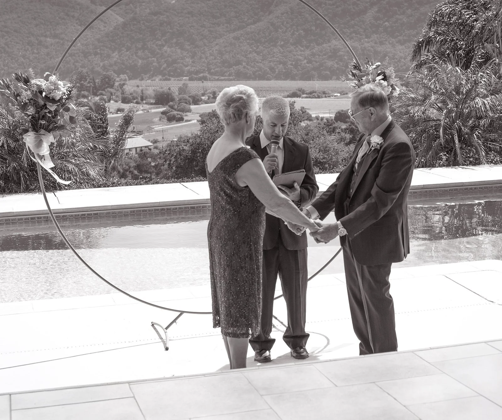 A black and white photo of a wedding ceremony with a couple holding hands and a person reading vows or giving a speech, outdoors with scenic hills in the background and floral decorations.
