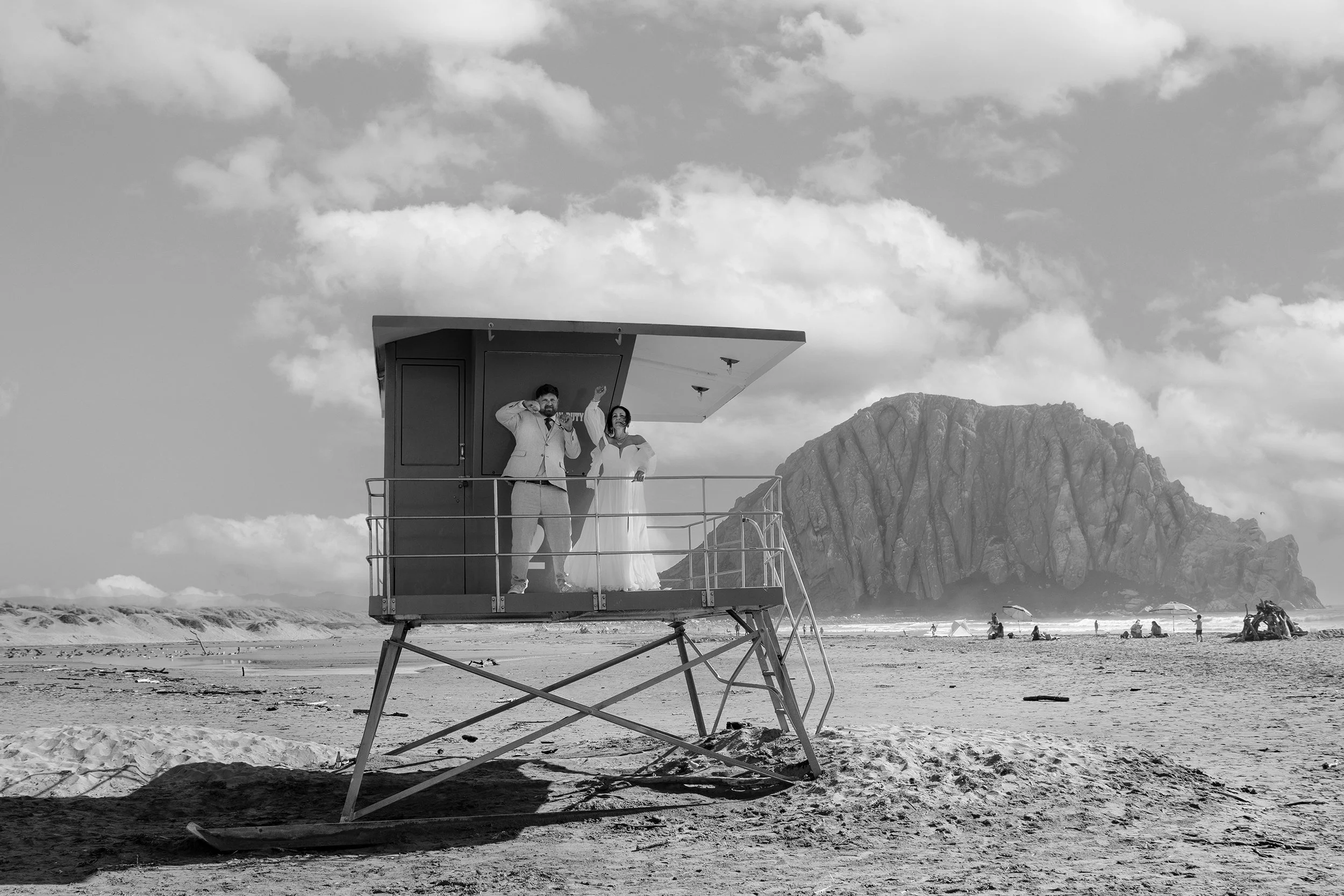 A couple dressed in wedding attire standing on a lifeguard tower on a beach with mountains in the background.