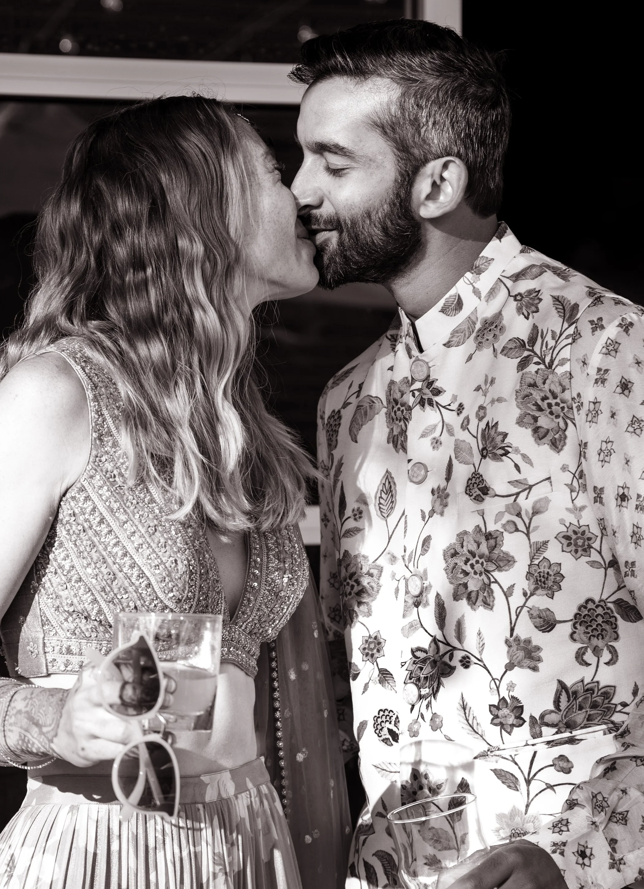 A couple about to kiss at a celebration, with woman holding a champagne glass and wearing a fancy dress, man wearing a floral shirt, in black and white.