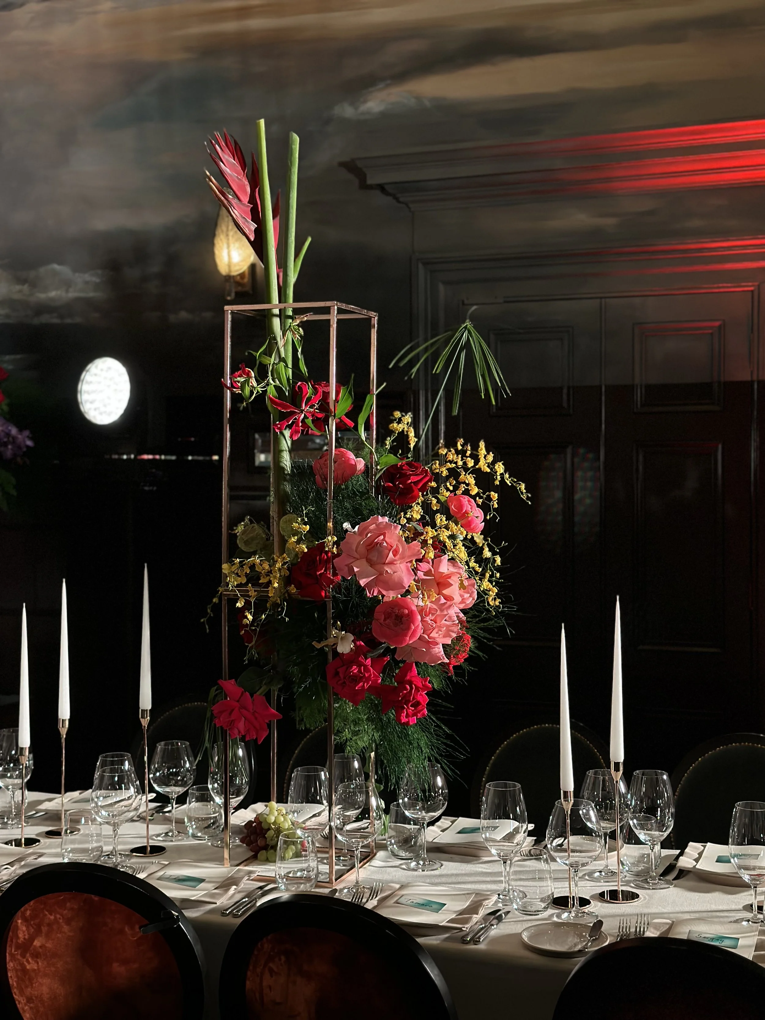 Elegant banquet table with a tall floral centerpiece consisting of pink and red flowers and greenery, set with wine glasses, plates, cutlery, and tall white candles, in a dimly lit room with dark walls and ambient lighting.