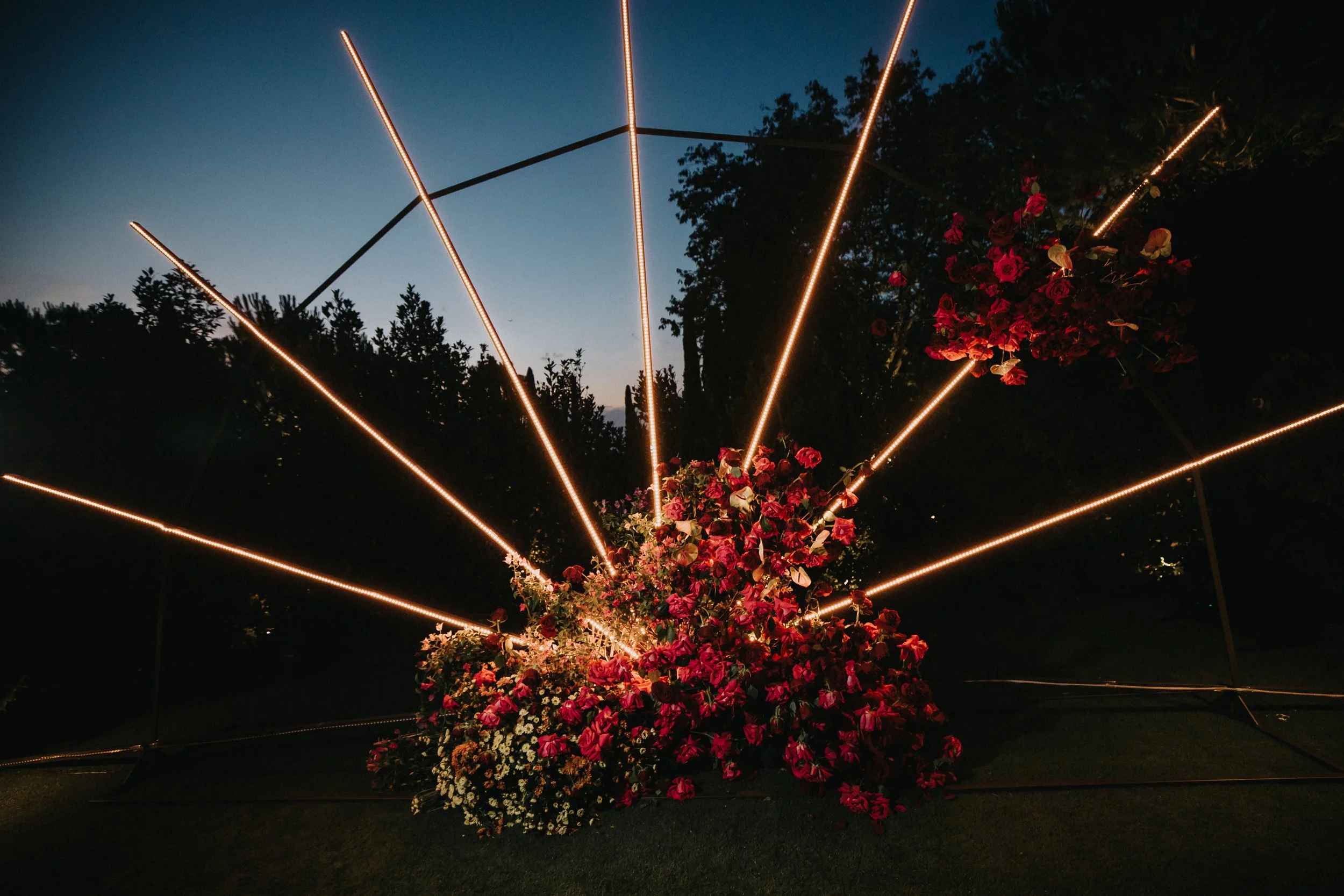 A floral arrangement with pink and white flowers illuminated by string lights in a dark outdoor setting at dusk.