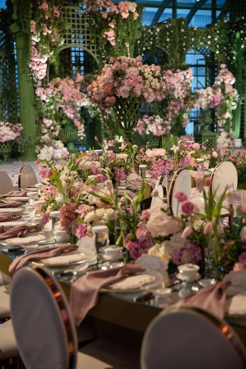 A wedding or event table decorated with pink and white flowers, with a large floral arrangement as a centerpiece in a tall green vase. The table is set with plates, napkins, and silverware, and surrounded by white chairs. The background features an ornate green and pink floral backdrop with greenery and flowers.