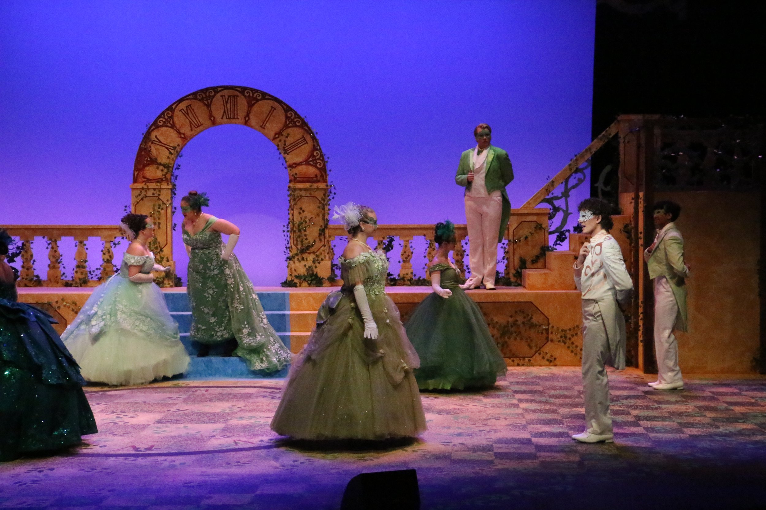 The stage scene depicts a theatrical performance with characters dressed in Victorian-style costumes, some wearing masks. The background features a blue backdrop with a large, ornate clock design. The set includes stairs and decorative elements suggesting a historical or fantasy setting.