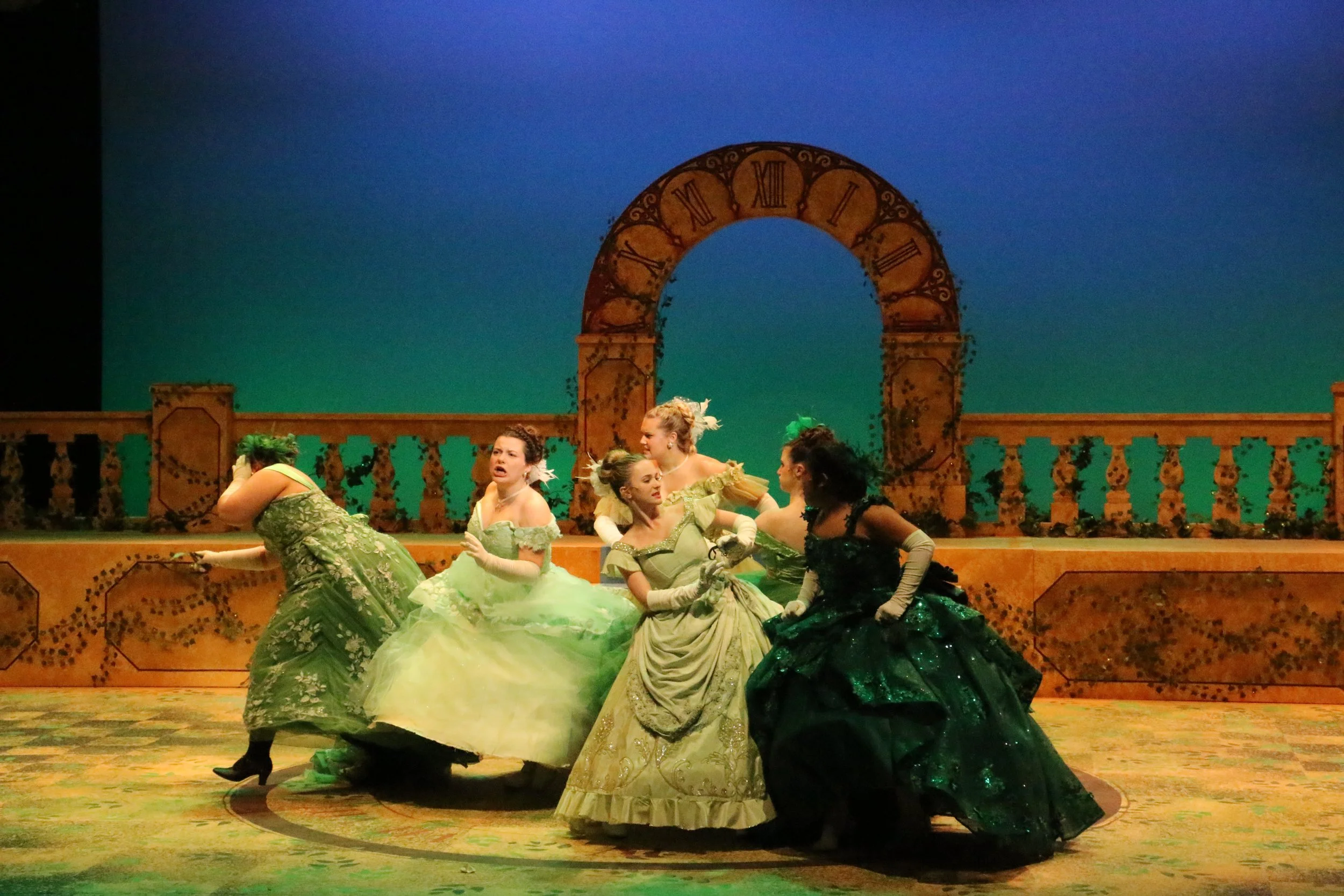 A scene from a theatrical performance featuring women in elaborate, colorful period costumes, sitting and conversing on stage with a backdrop resembling a garden or outdoor setting with an arch and decorative railing.