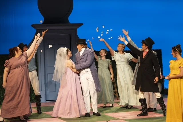 A theatrical stage scene with actors in period costumes, including a bride and groom, celebrating by throwing confetti or paper pieces into the air.